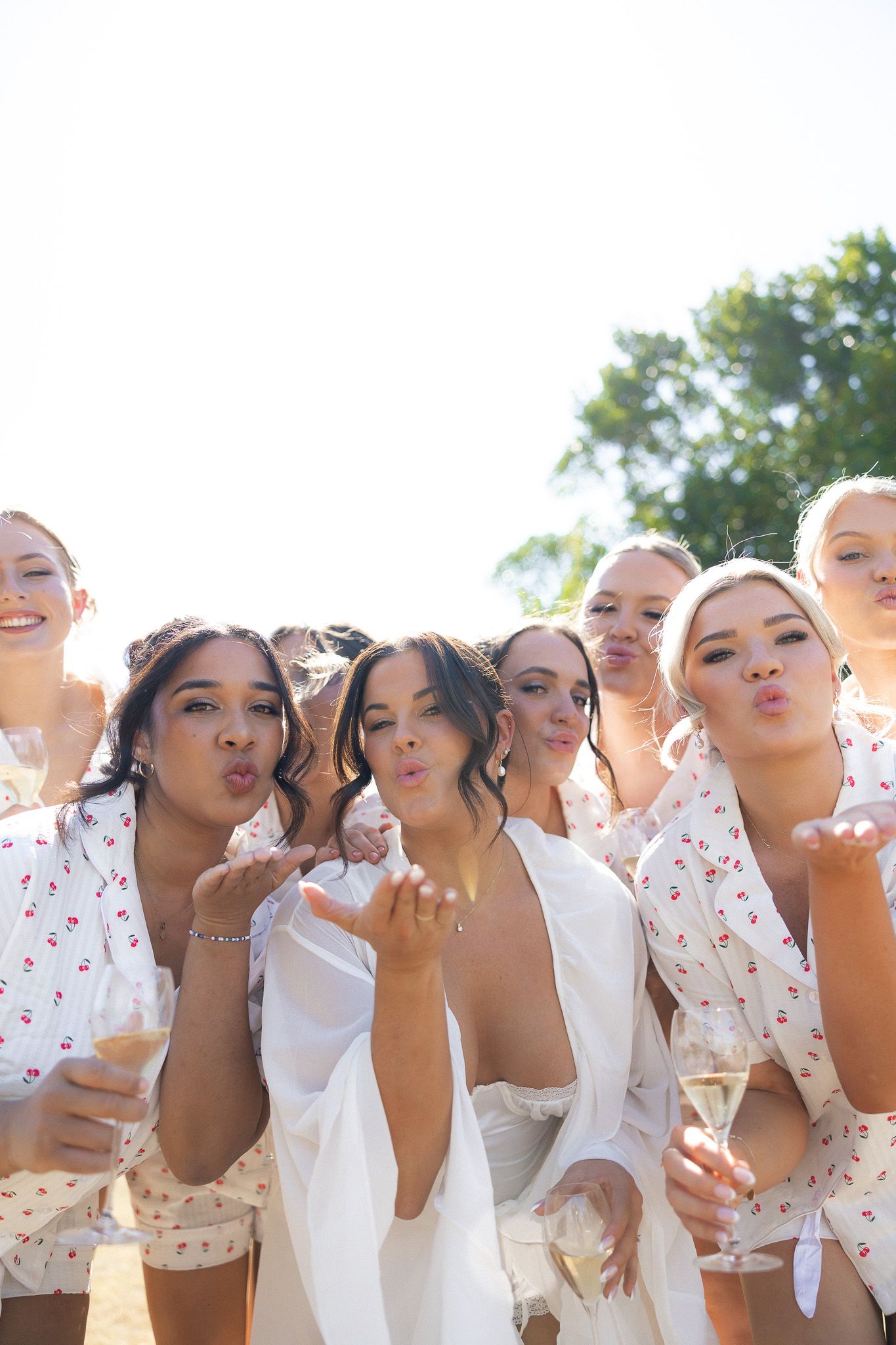 Bride and bridesmaids in robes blowing kisses while holding champagne glasses outdoors.