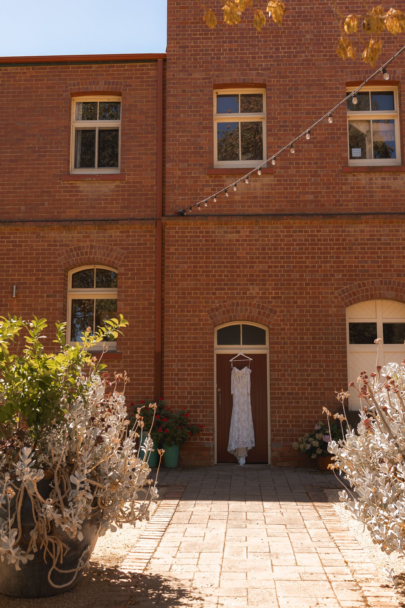 White wedding dress hanging on a brick building door in a sunlit courtyard with string lights and lush greenery.