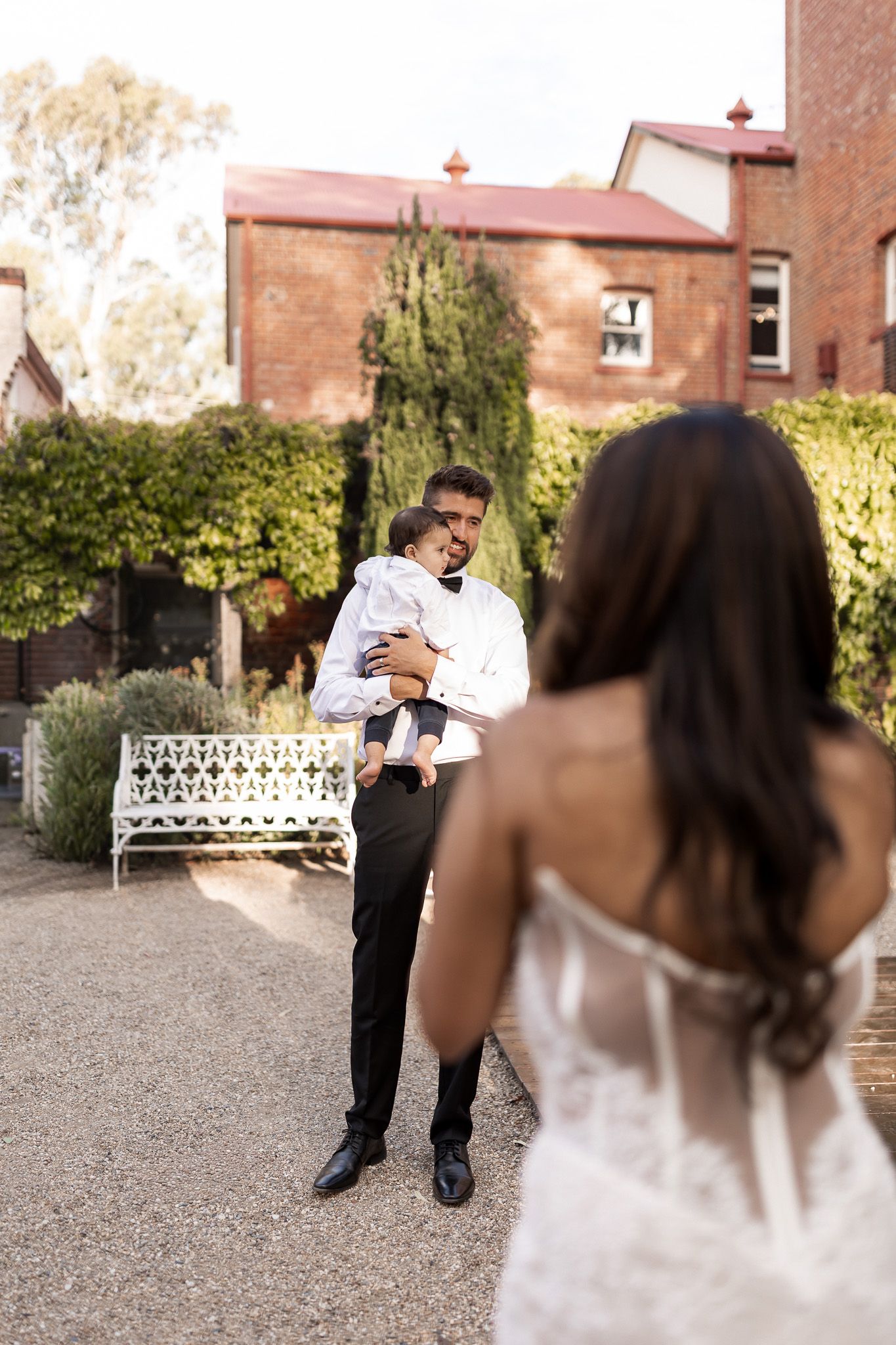 Groom in white shirt holding a baby while bride in lace dress stands in foreground at outdoor venue.