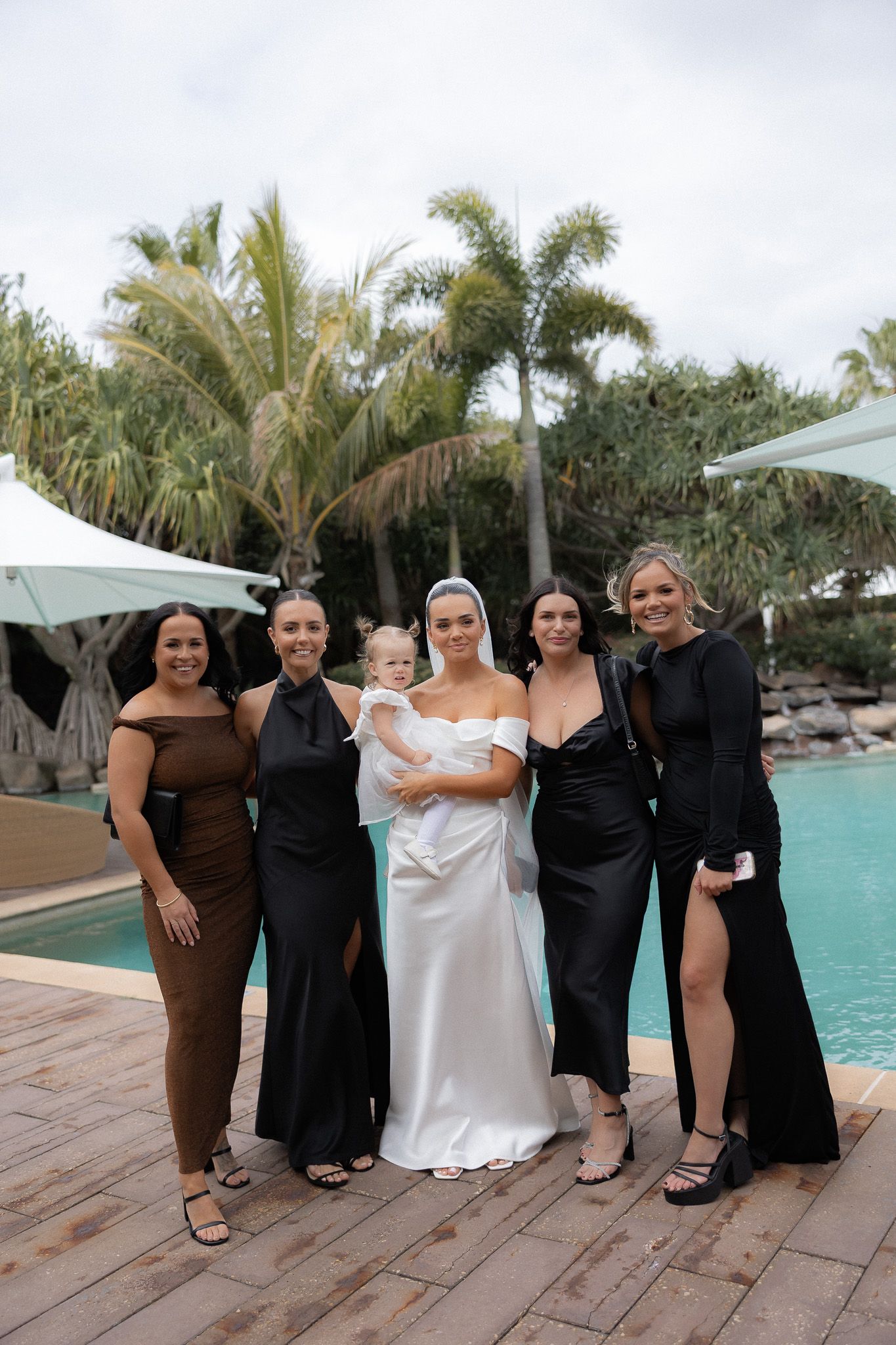 Bride in white gown holds baby surrounded by bridesmaids in black dresses beside a pool with palm trees.