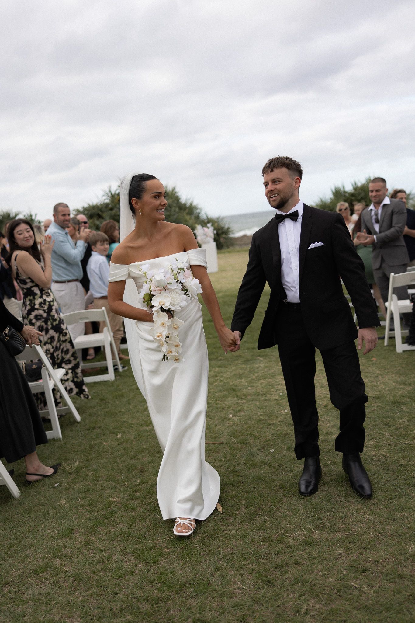 Smiling bride in off-shoulder white gown and groom in tuxedo walk down aisle after ceremony.