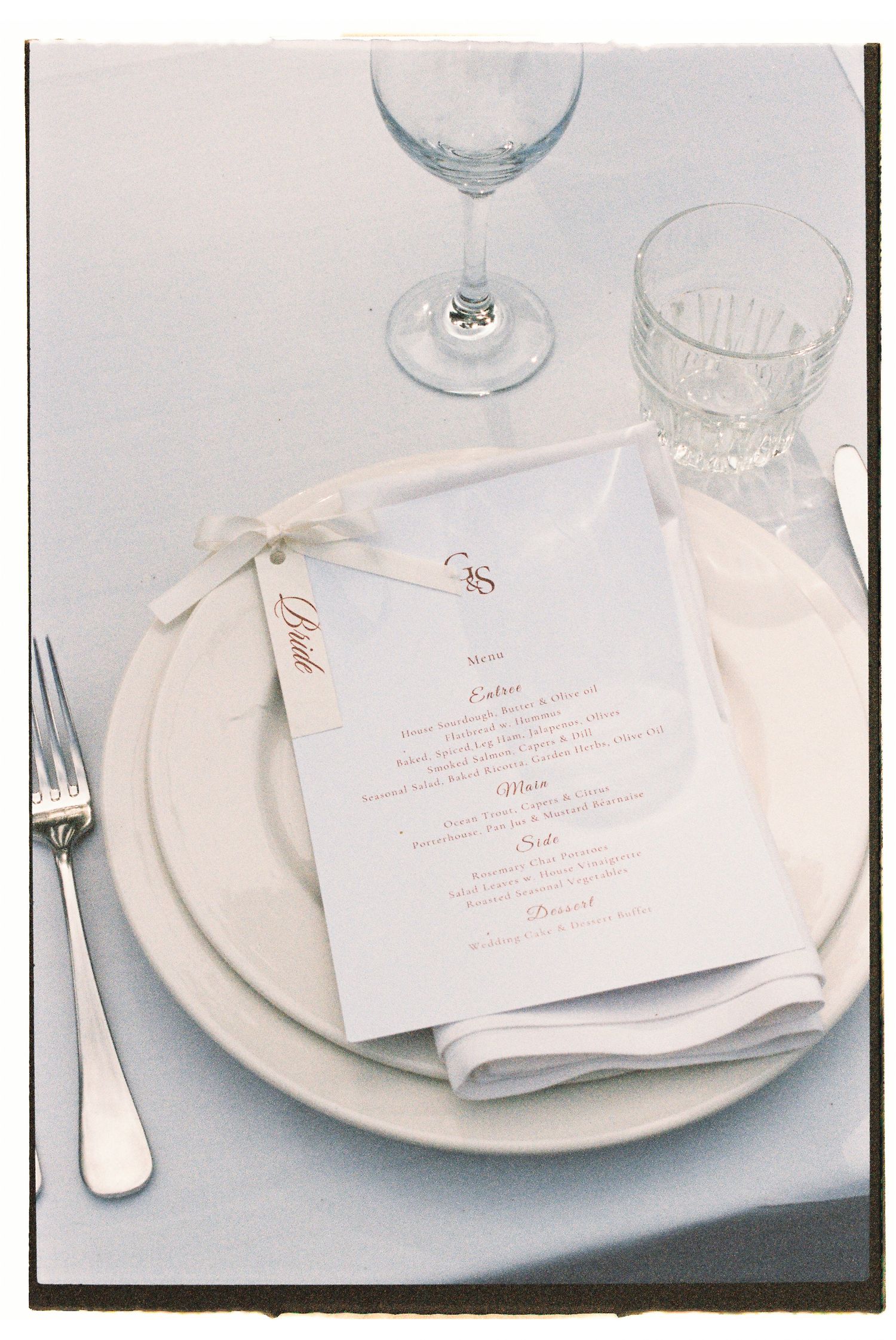 Elegant wedding place setting with white plates, menu card, folded napkin, fork, and crystal glassware on a white tablecloth.