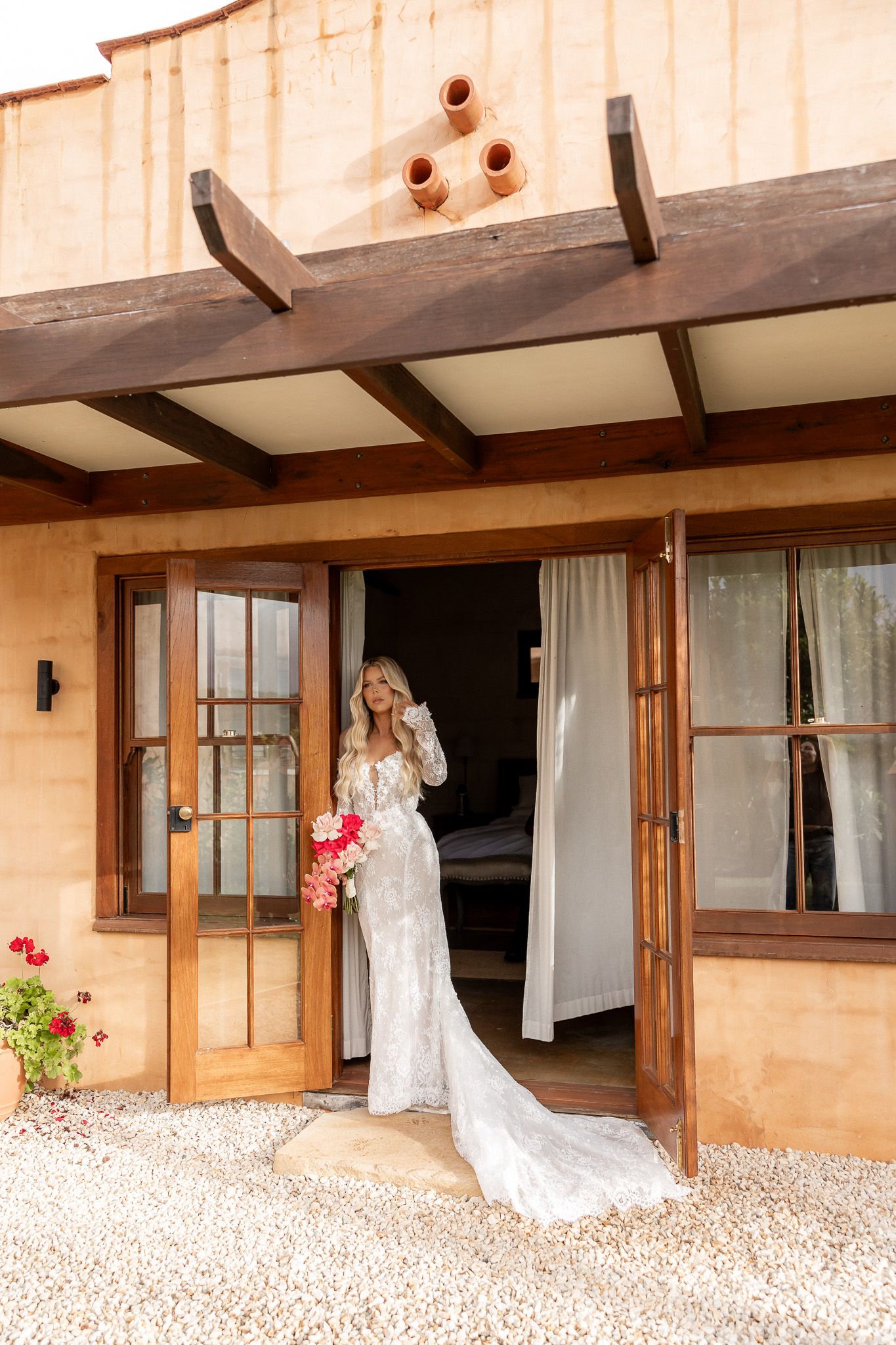 Bride in lace wedding gown with train holds pink bouquet standing in doorway of rustic adobe building.