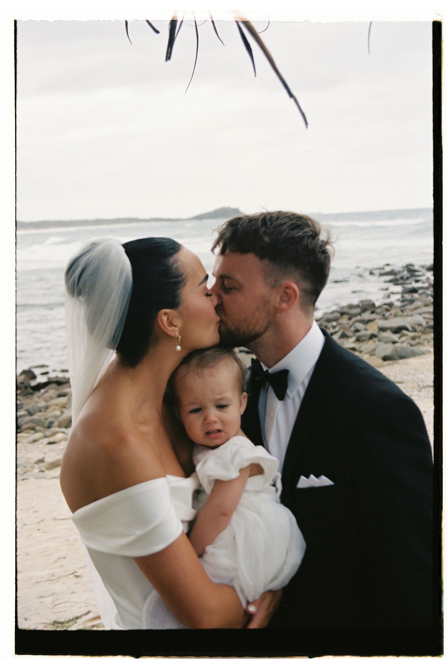 Newlywed couple kissing while holding infant on beach, groom in black tuxedo.