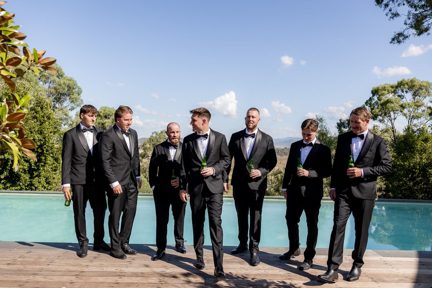 Six groomsmen in black tuxedos standing by a pool on a sunny wedding day.