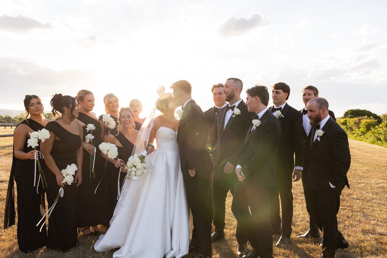 Wedding party in black attire surrounds kissing bride and groom in golden sunset light on open field.