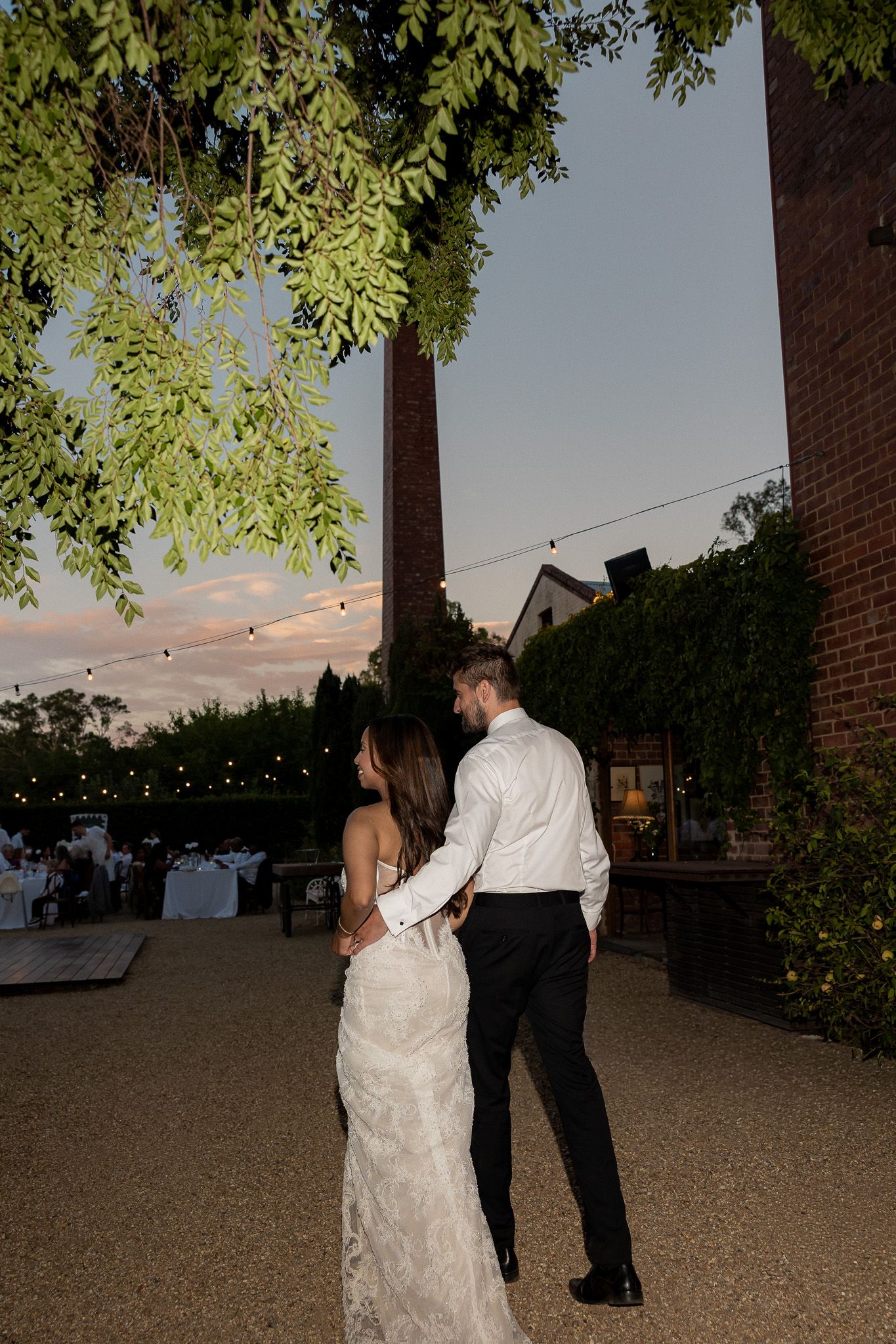 Bride and groom share a romantic moment outdoors at dusk, surrounded by string lights and lush greenery at a wedding venue.