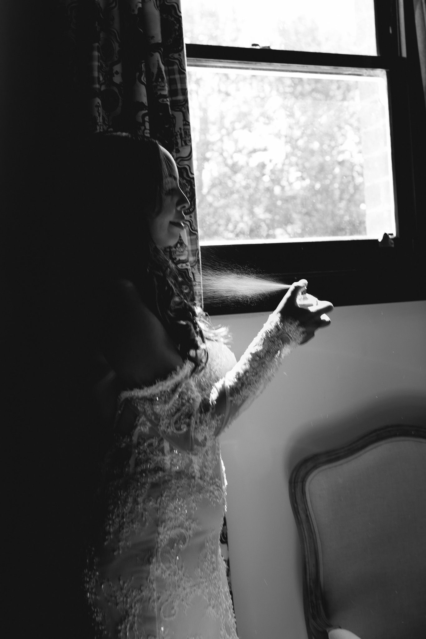 Bride in lace wedding dress spraying perfume by a sunlit window in a dark room.