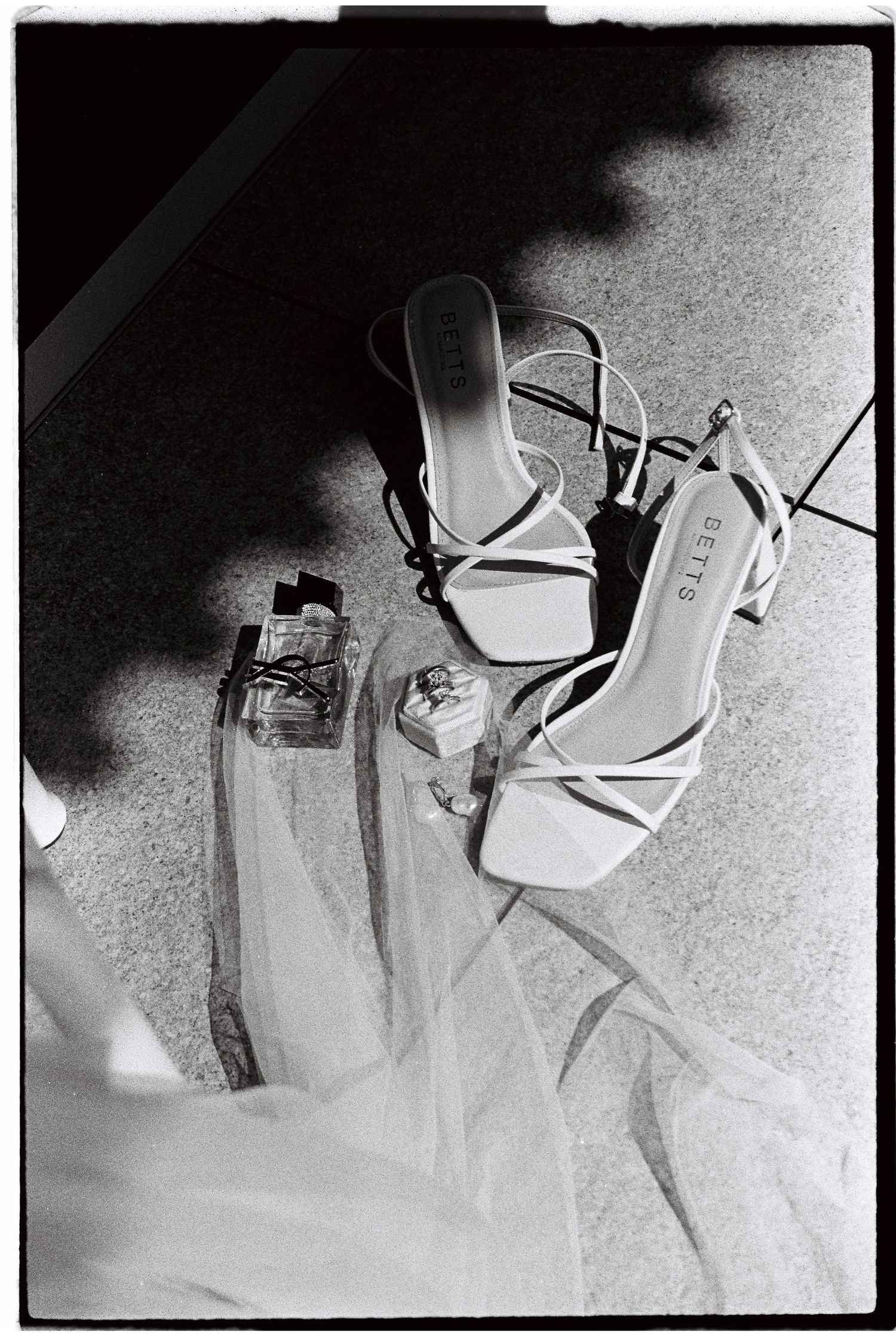 White strappy Betts heels with perfume bottles on floor in black and white wedding flat lay.