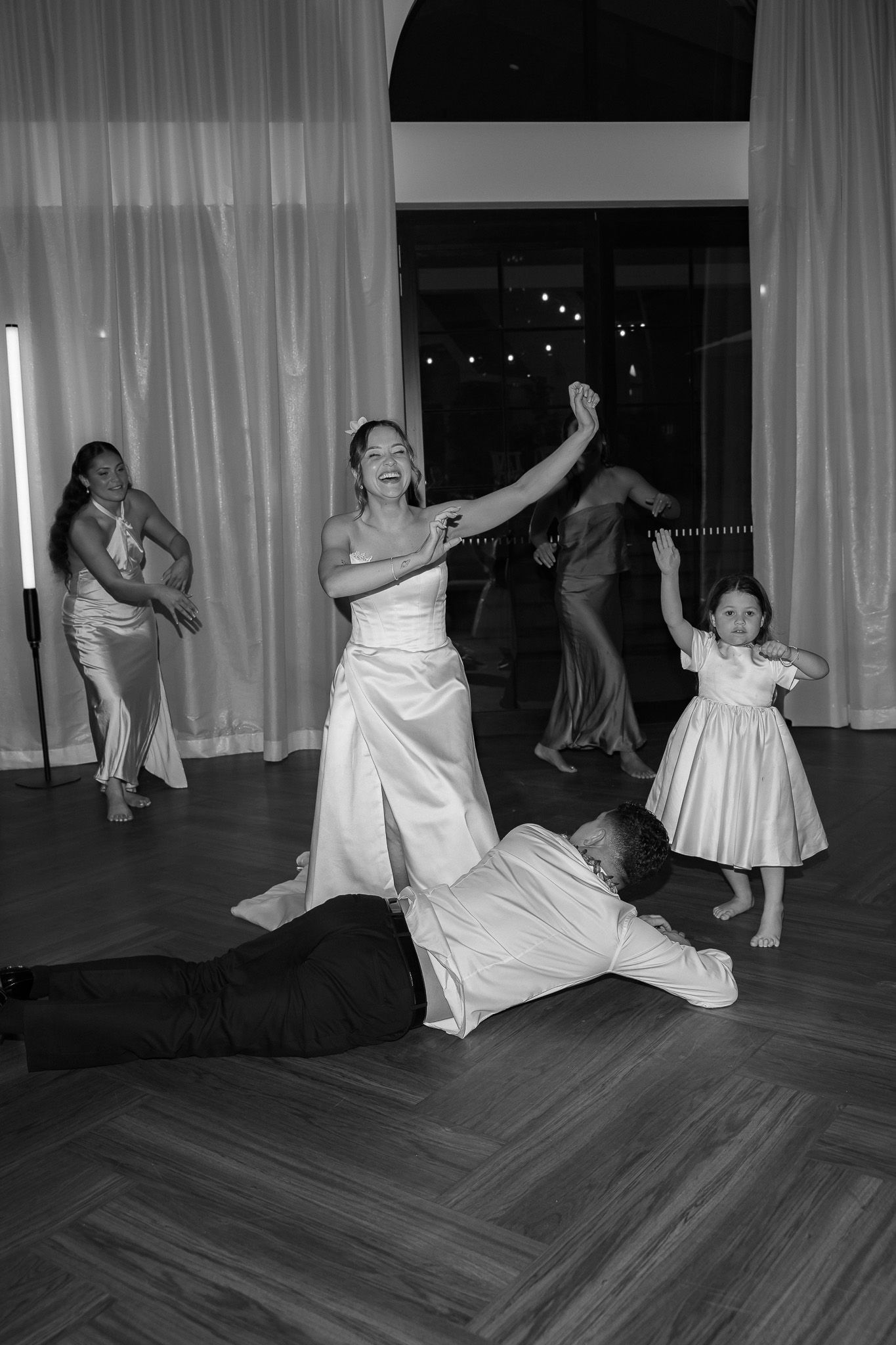 Black and white reception photo of bride dancing with flower girl while groom lies on dance floor.