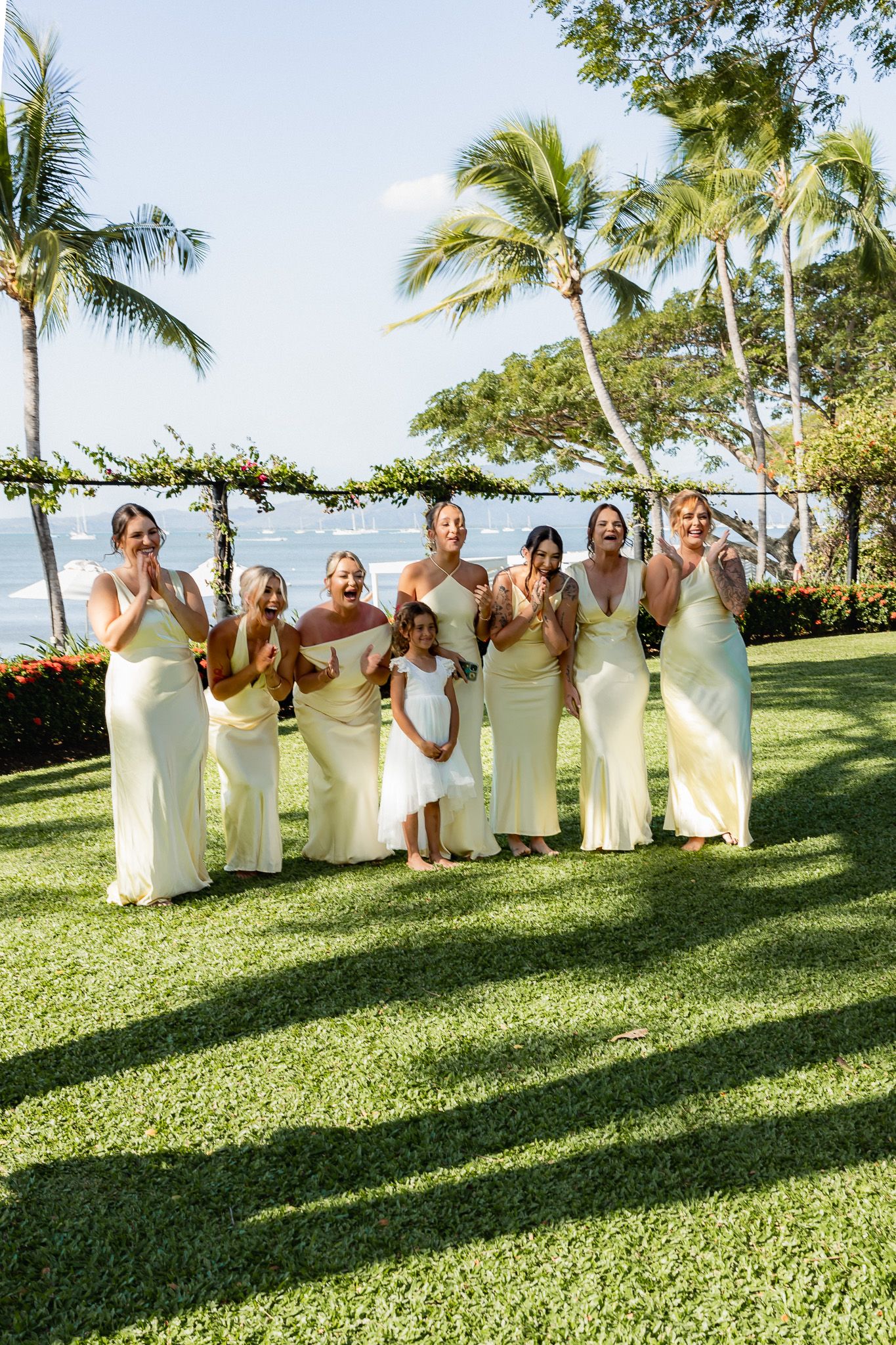 Bridesmaids in yellow dresses and flower girl clapping joyfully at beachside wedding ceremony with palm trees.