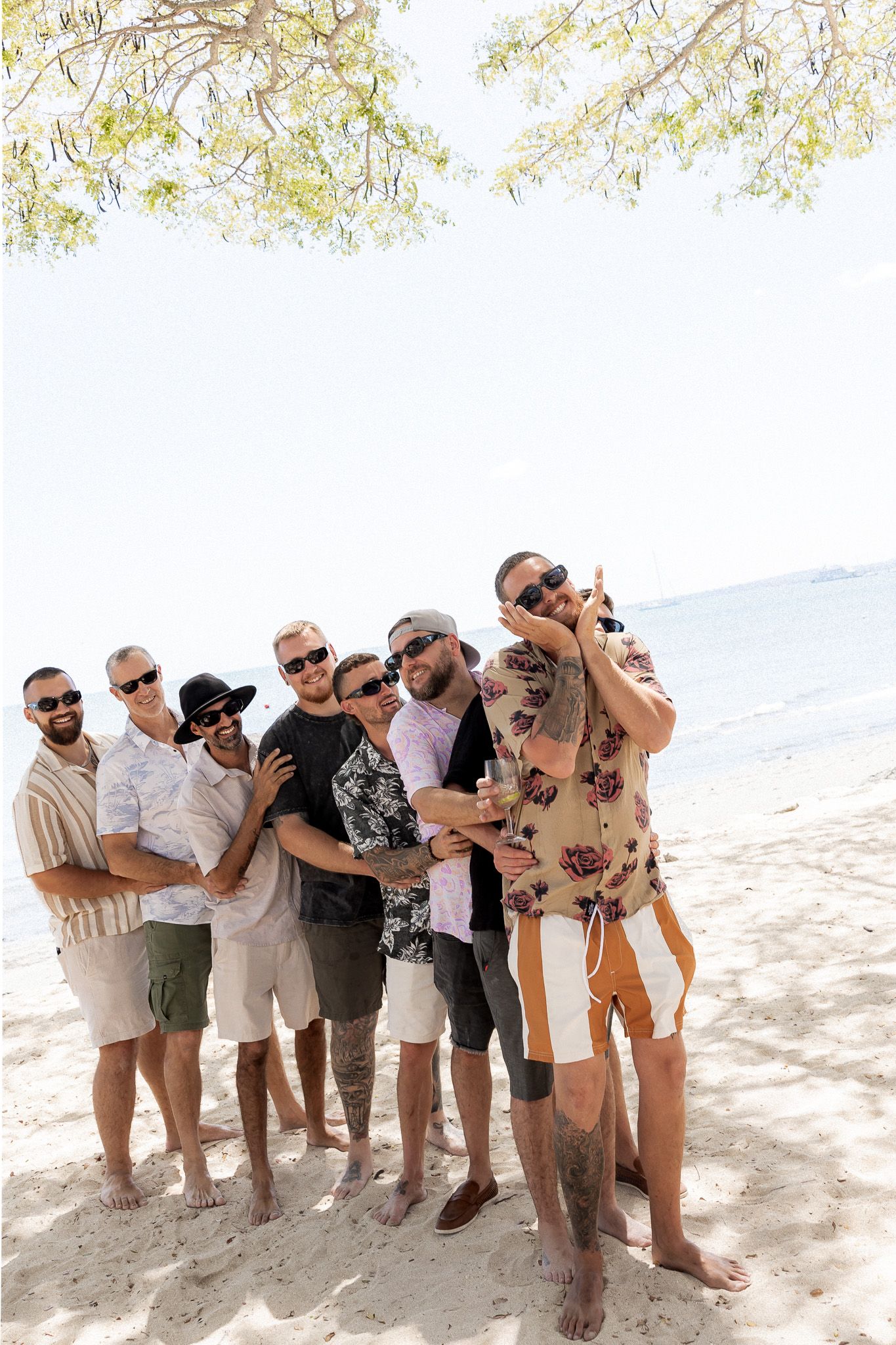 Group of men in sunglasses and casual summer outfits posing together on a sunny sandy beach near the ocean.