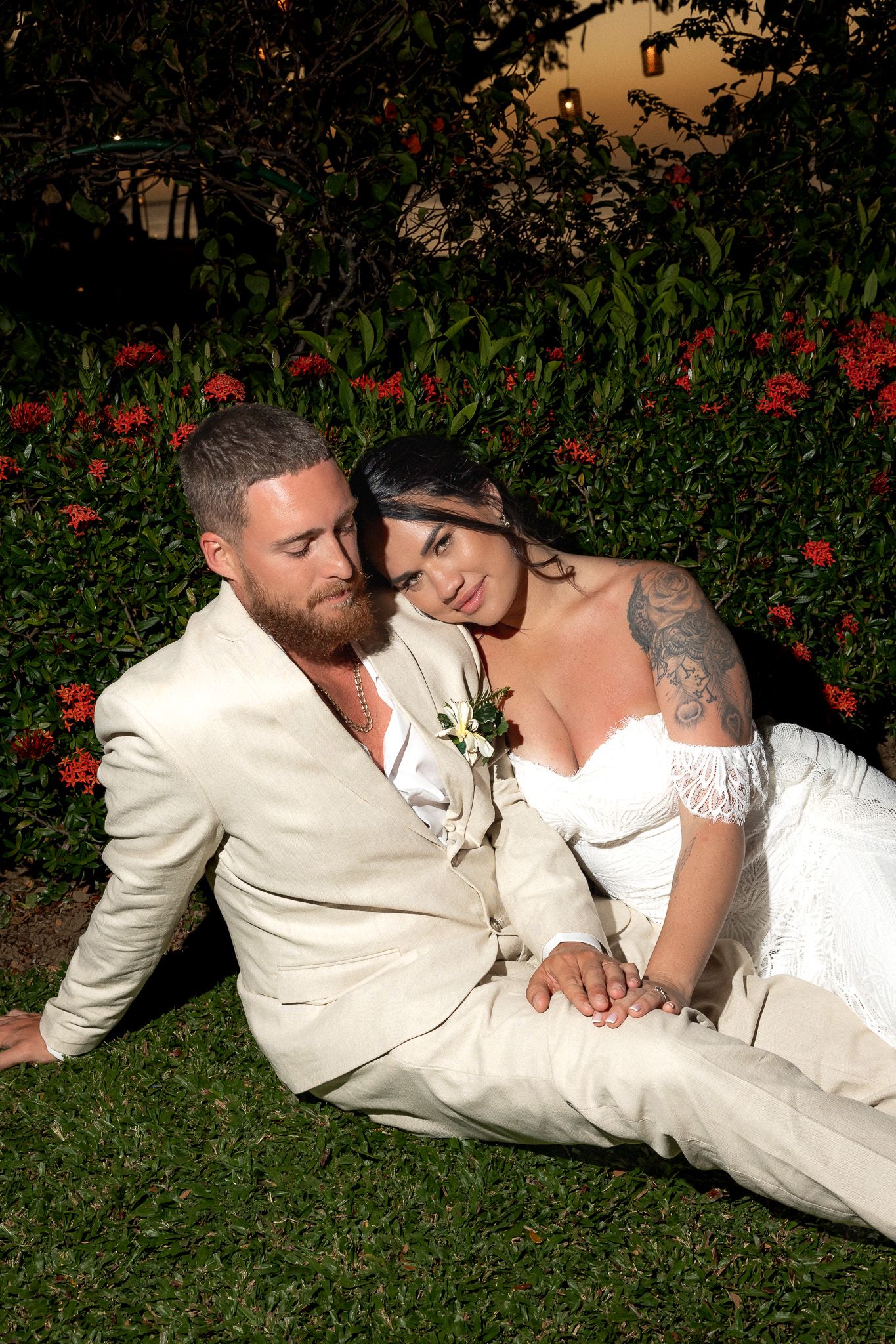 Bride and groom in wedding attire sit together on grass surrounded by red tropical flowers.
