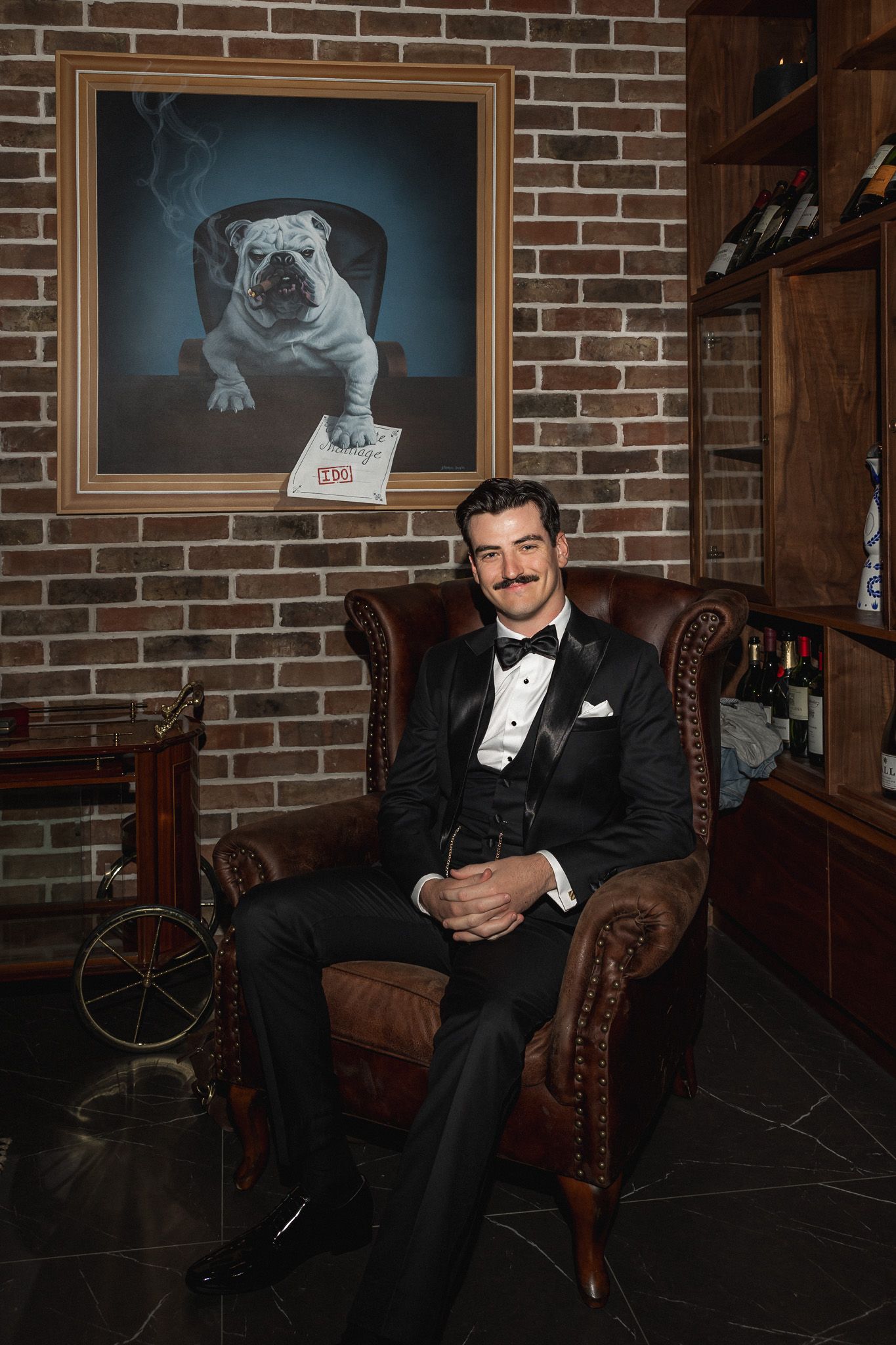 Dapper groom in black tuxedo sitting in leather chair holding champagne in elegant cigar lounge.