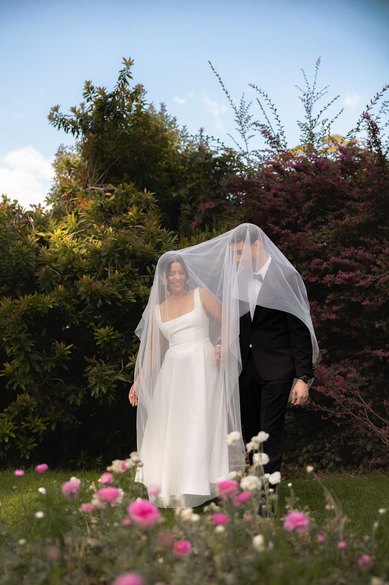 Bride and groom share a romantic moment under a white veil in a garden with pink flowers and lush greenery.