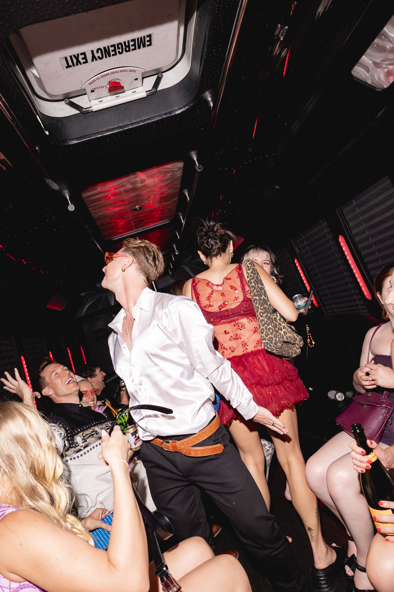 Young adults dancing and celebrating on a party bus with red lights, drinks, and an emergency exit sign overhead.