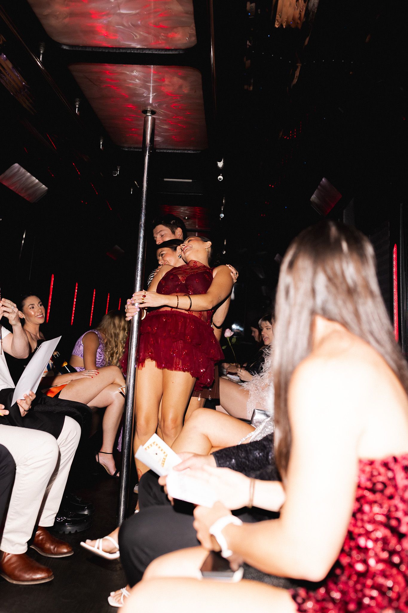 Two women in red dresses embrace near a pole in a lively nightclub with seated guests around them.
