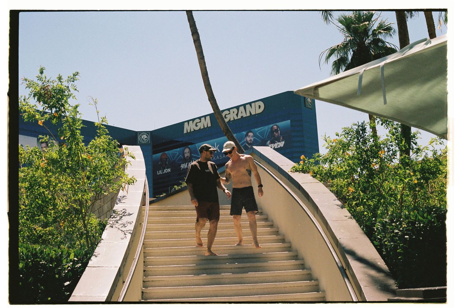 Two shirtless men walking up outdoor stairs at MGM Grand resort on a sunny day.