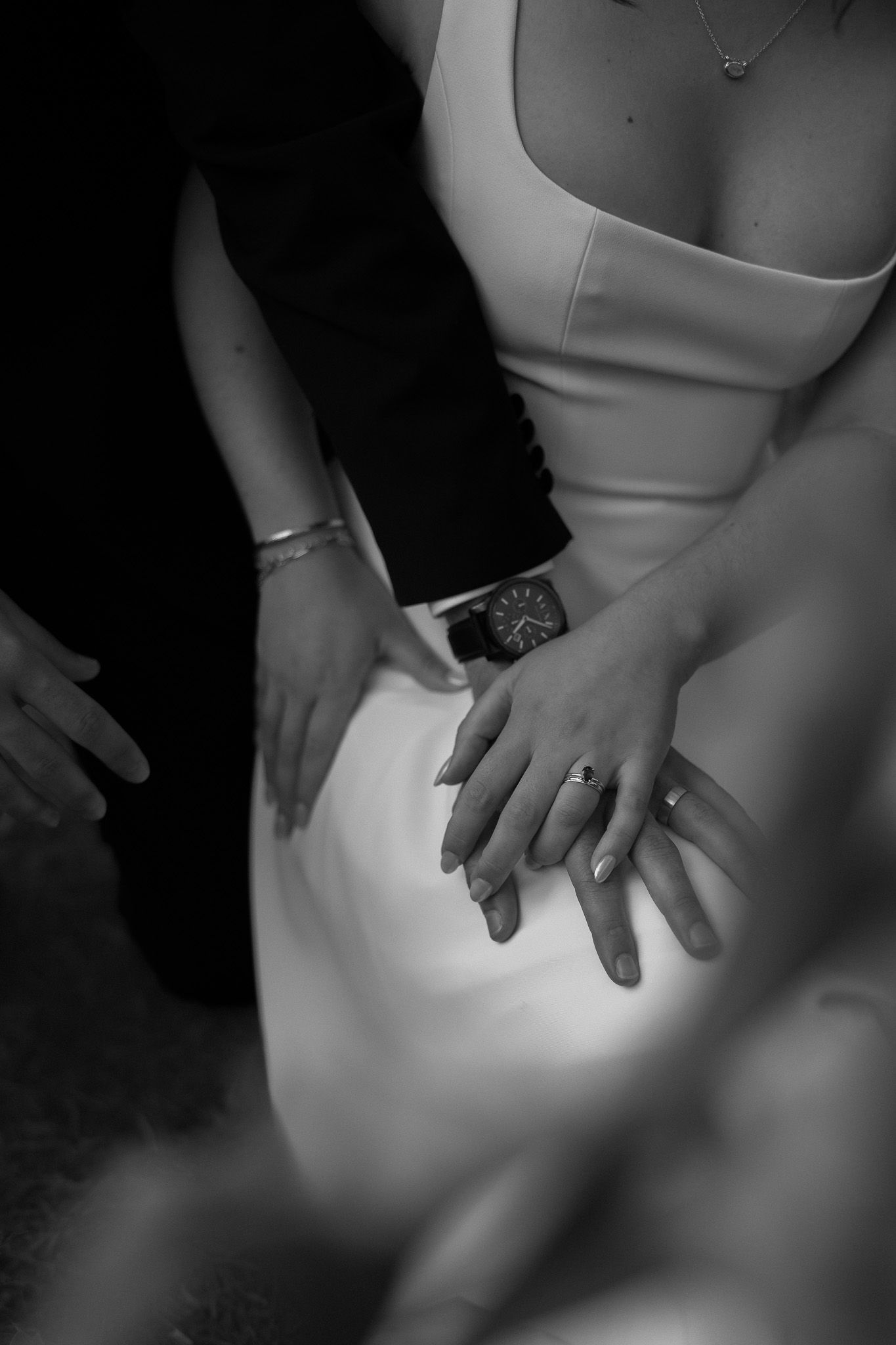 Black and white photo of bride and groom hands with wedding rings resting on white wedding dress.