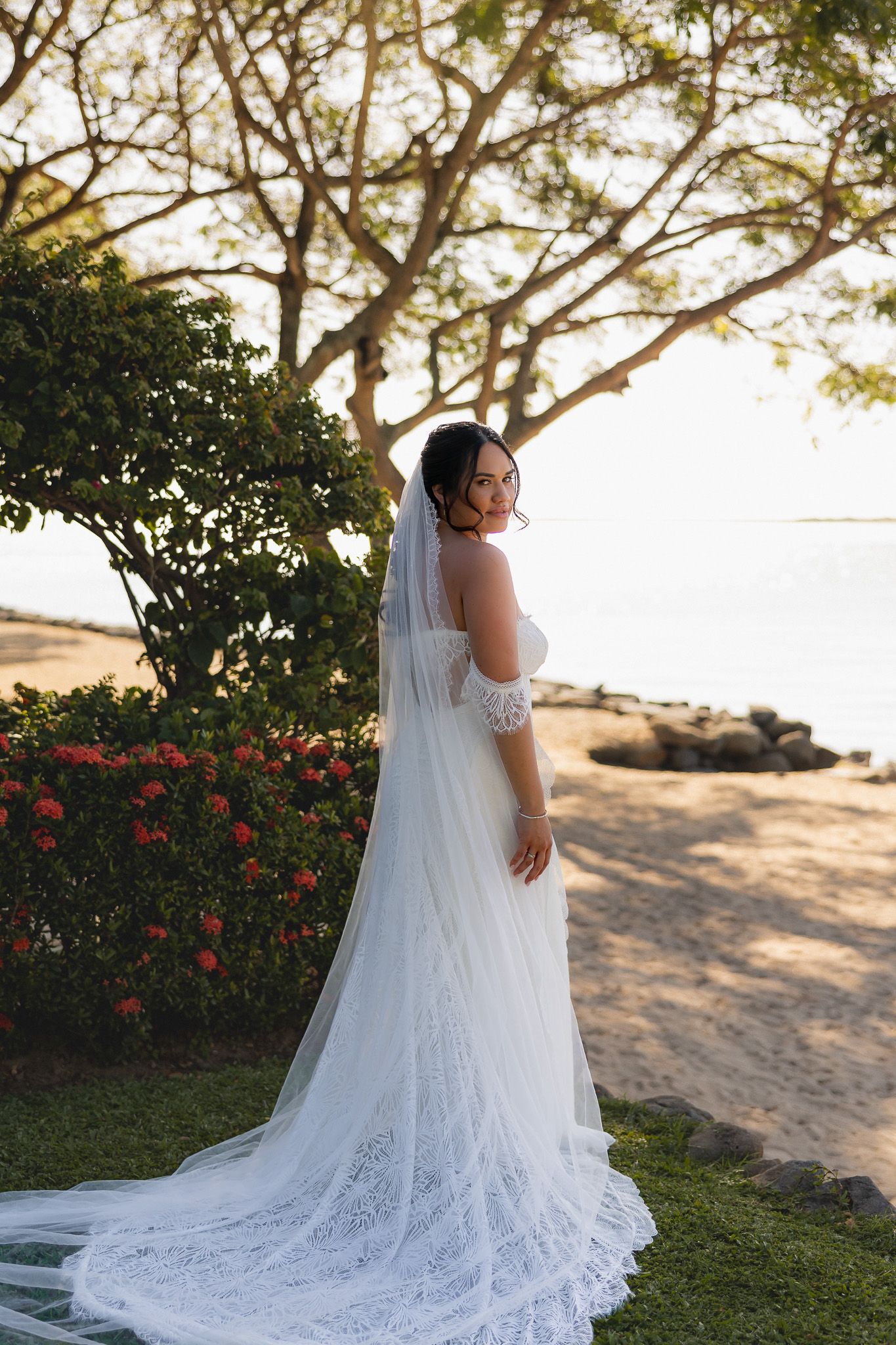 Bride in lace wedding gown and cathedral veil posing near tropical beach at sunset.