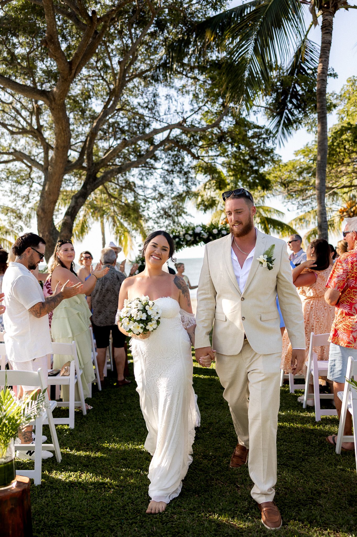 Newlywed couple walking hand in hand down aisle at tropical outdoor wedding as guests applaud.