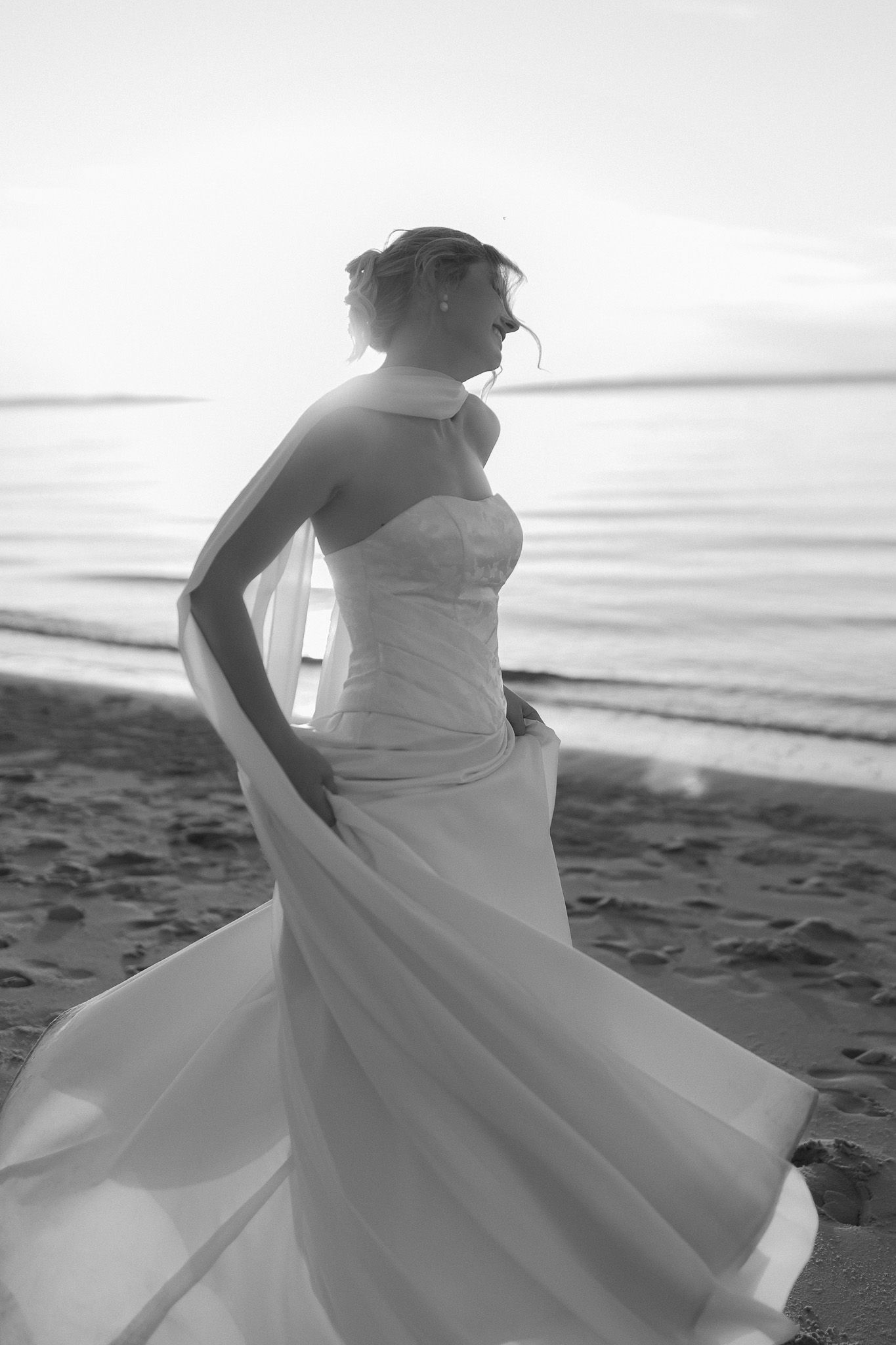 Black and white photo of a bride in a flowing strapless gown on a beach at sunset, her veil billowing in the wind.