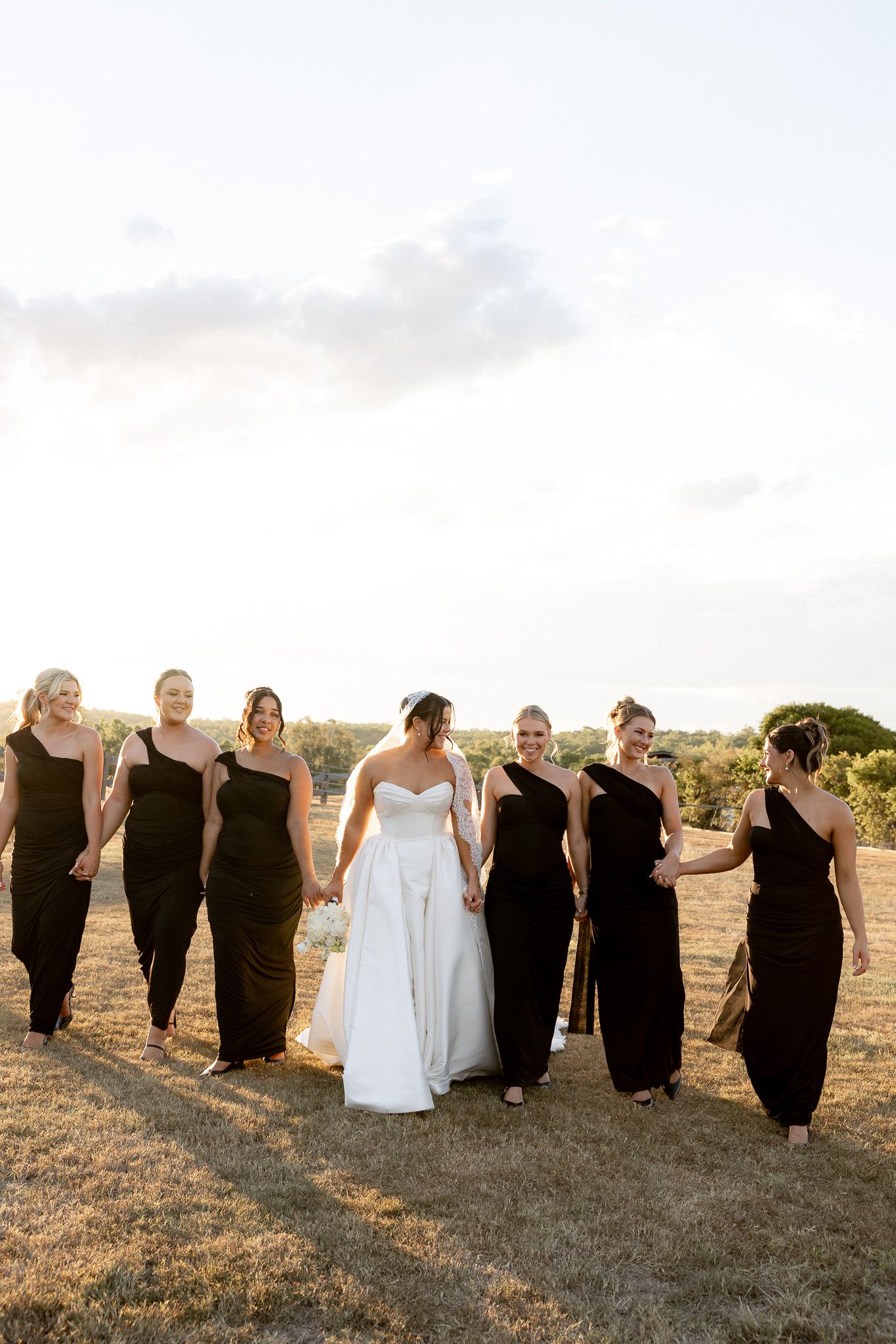 Bride in white strapless gown walks with bridesmaids in black dresses across a sunlit golden field.