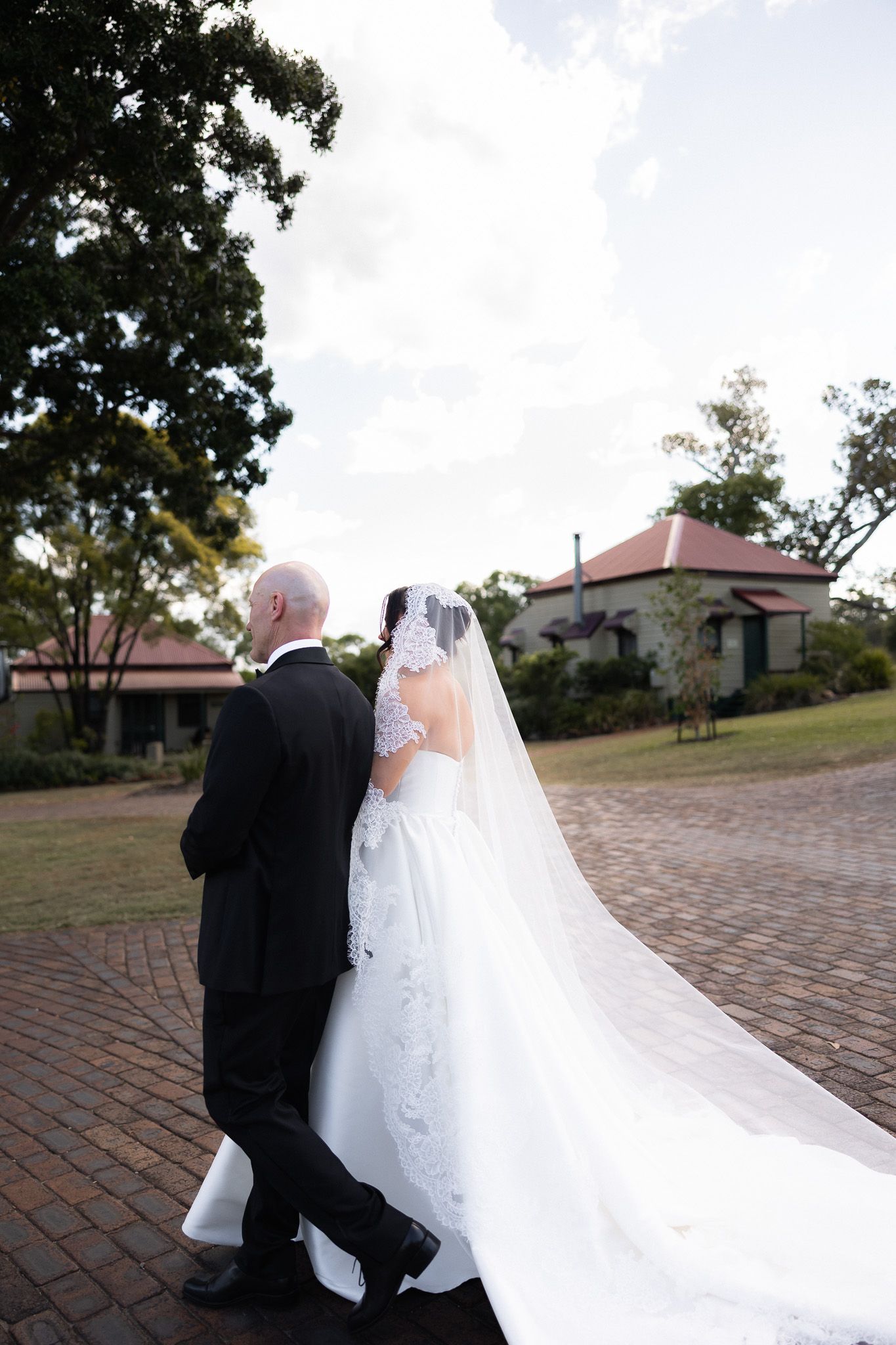 Bride in lace cathedral veil and gown walking with father along brick path to ceremony.
