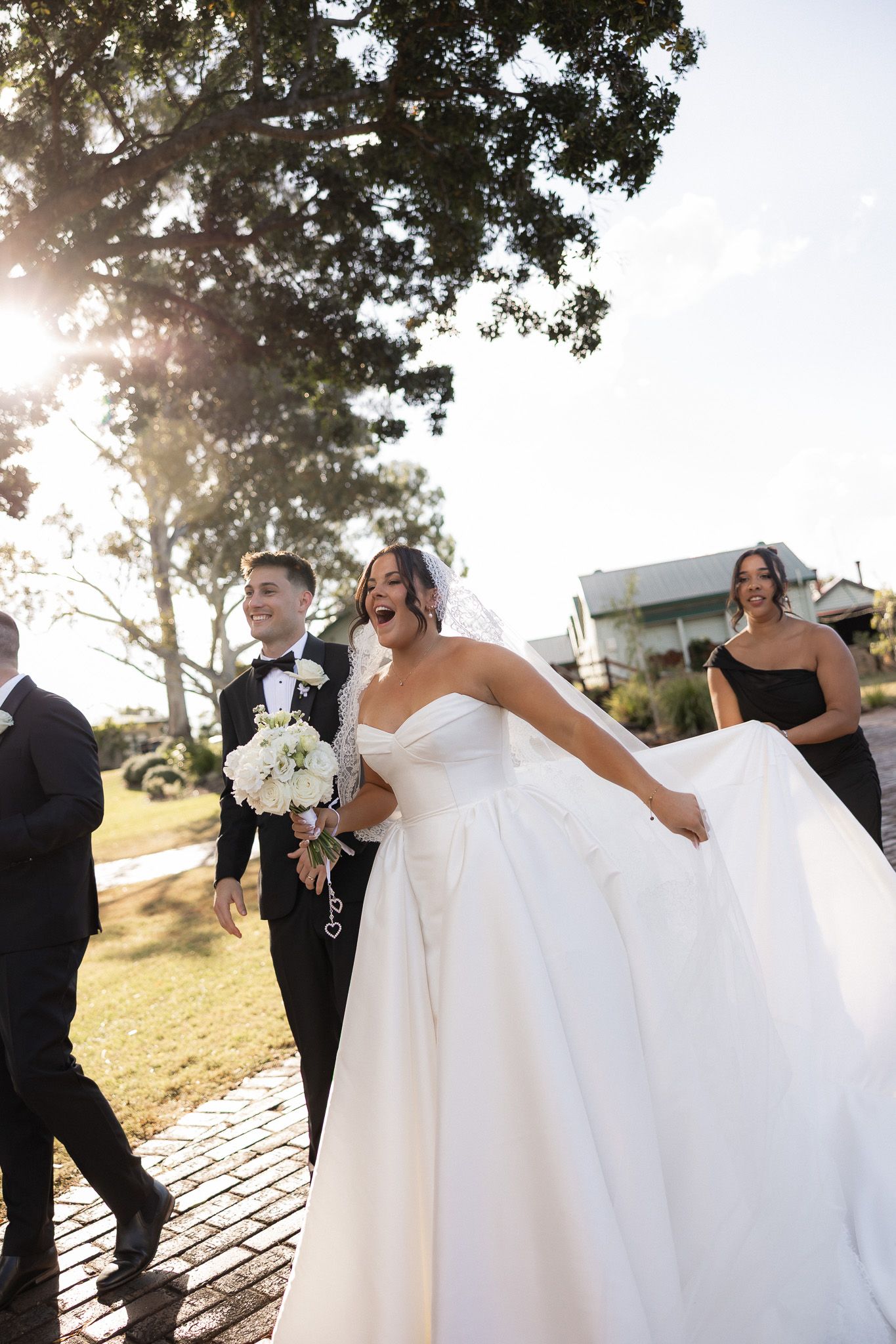Joyful bride in white ballgown laughing with groom in tuxedo and wedding party walking outdoors in golden sunlight.