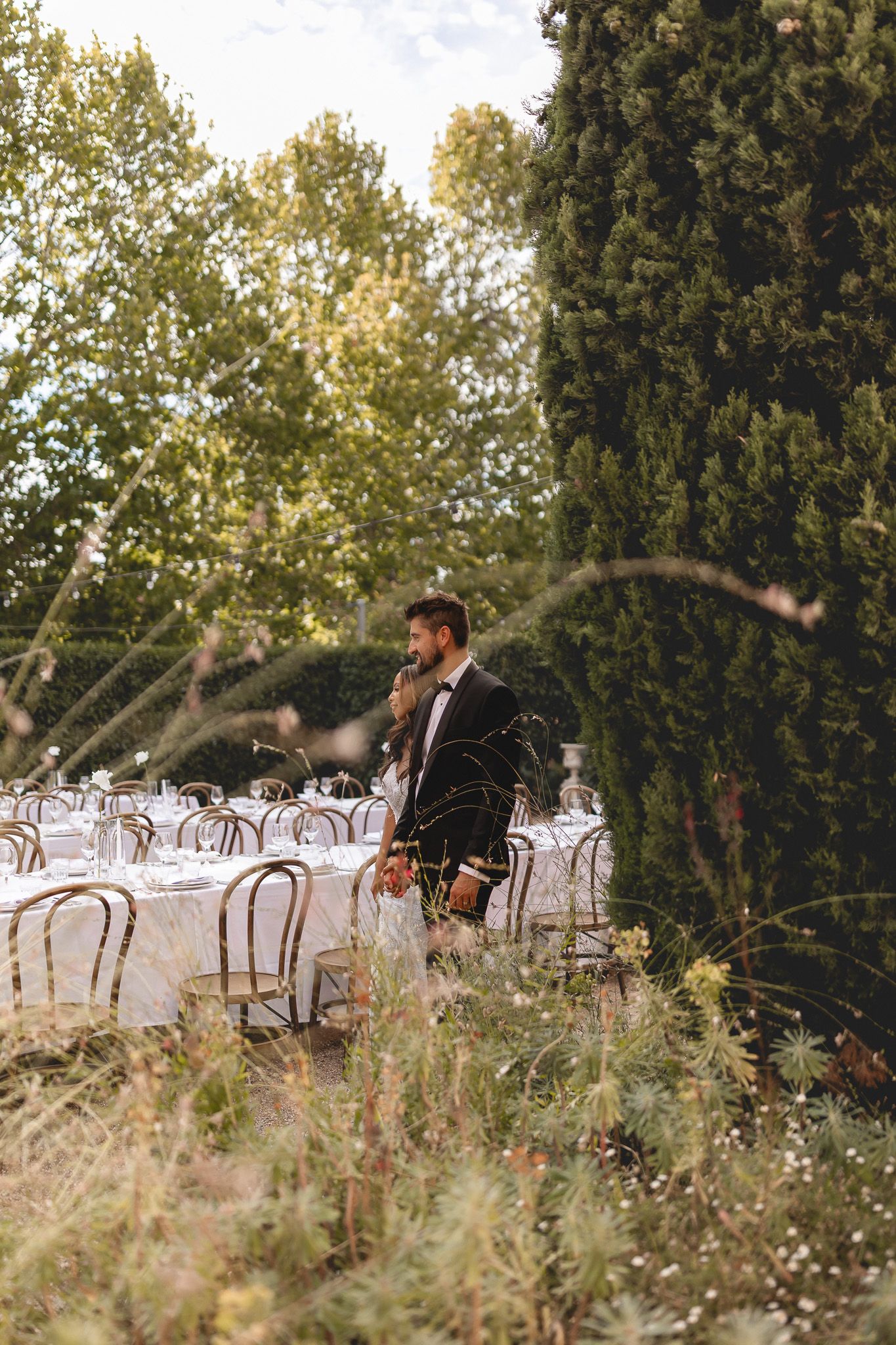 Groom in black tuxedo standing beside a long outdoor wedding reception table surrounded by lush cypress trees and greenery.