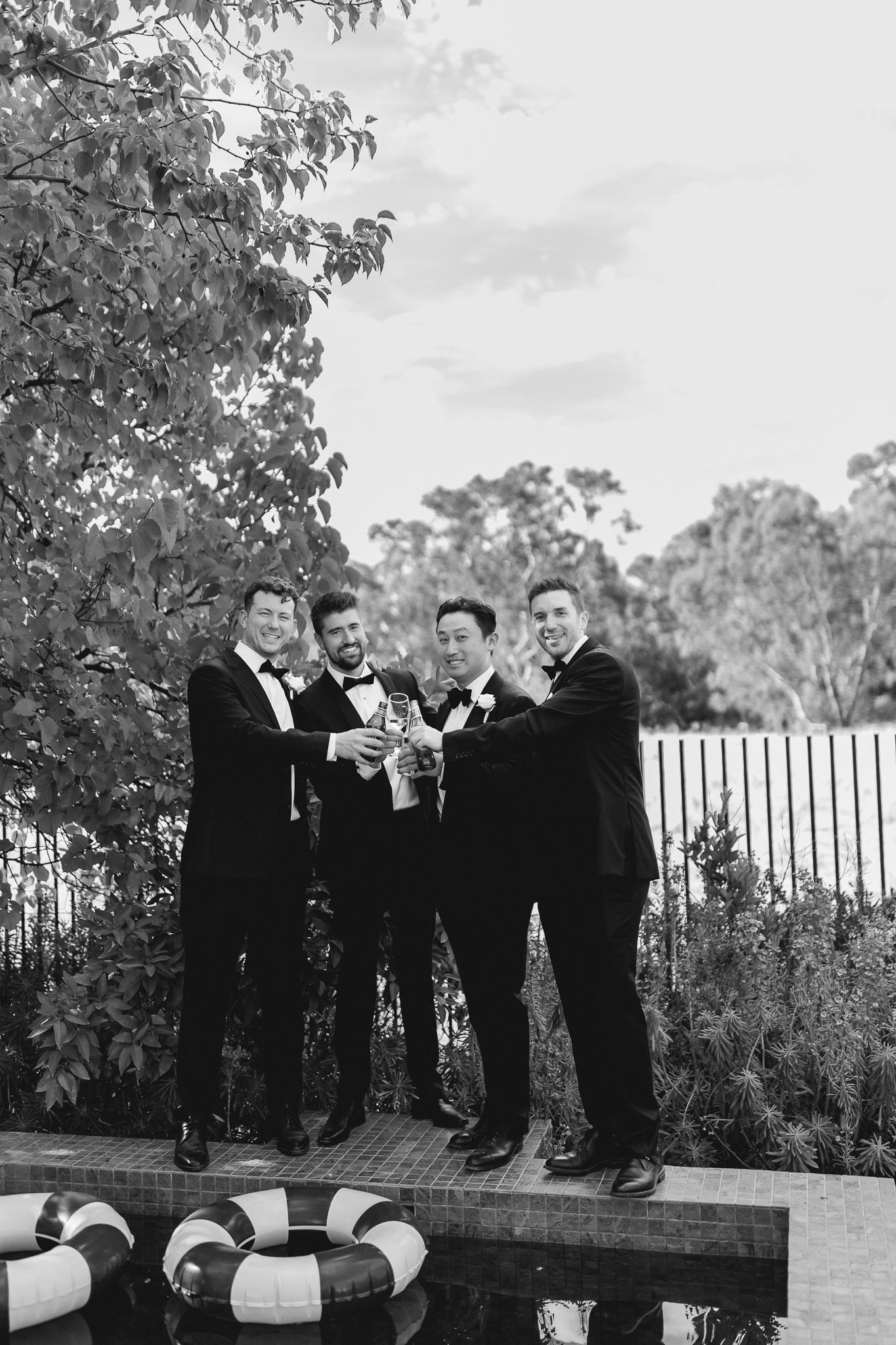 Four groomsmen in tuxedos toasting with champagne outdoors near a pool at a wedding celebration.