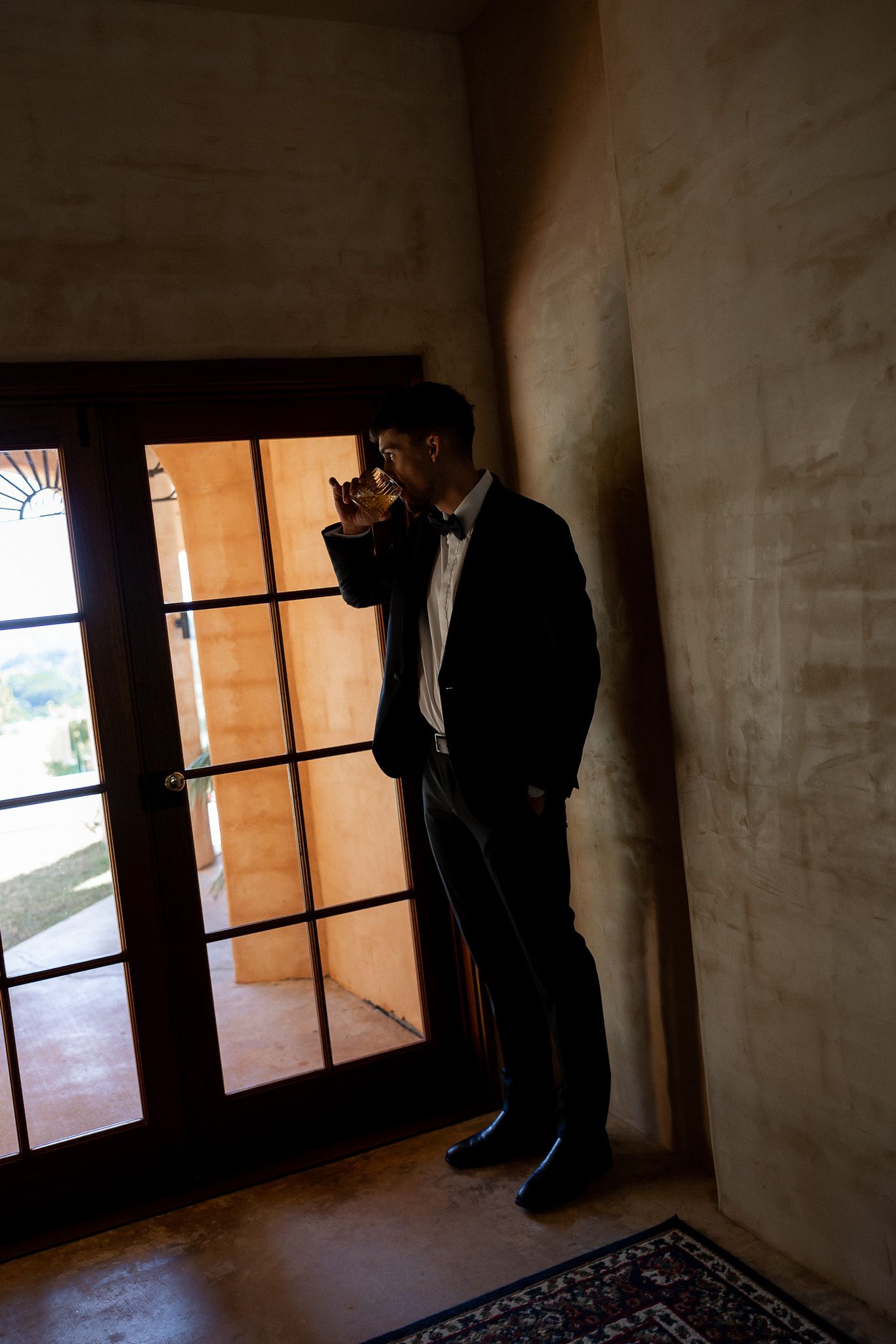 Elegant groom in black tuxedo drinking whiskey while standing by French doors in moody light.