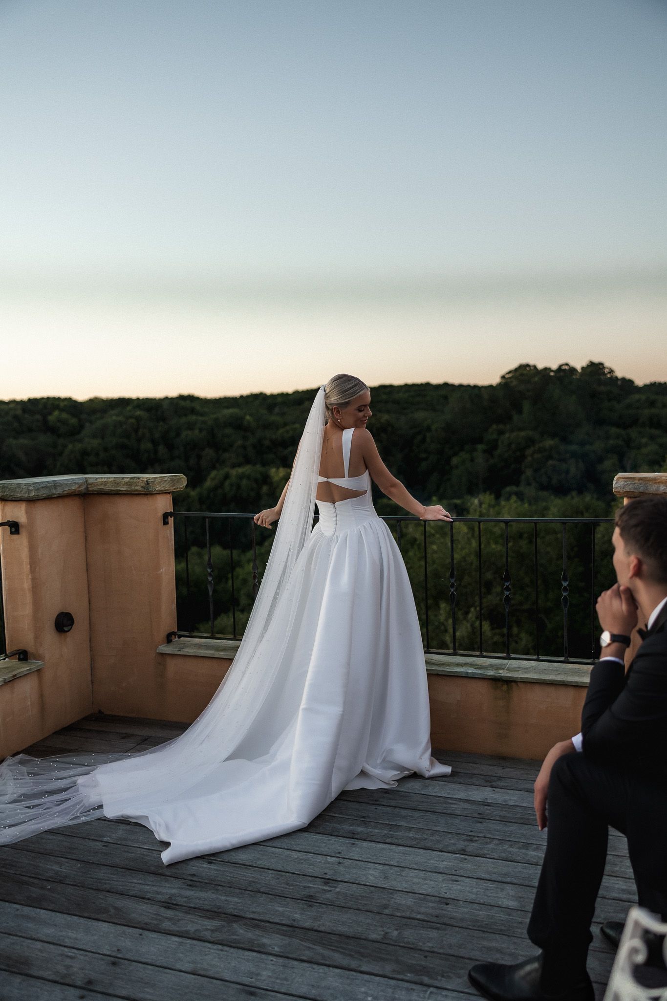 Bride in white gown and veil stands on a rooftop terrace at dusk with scenic forest views.