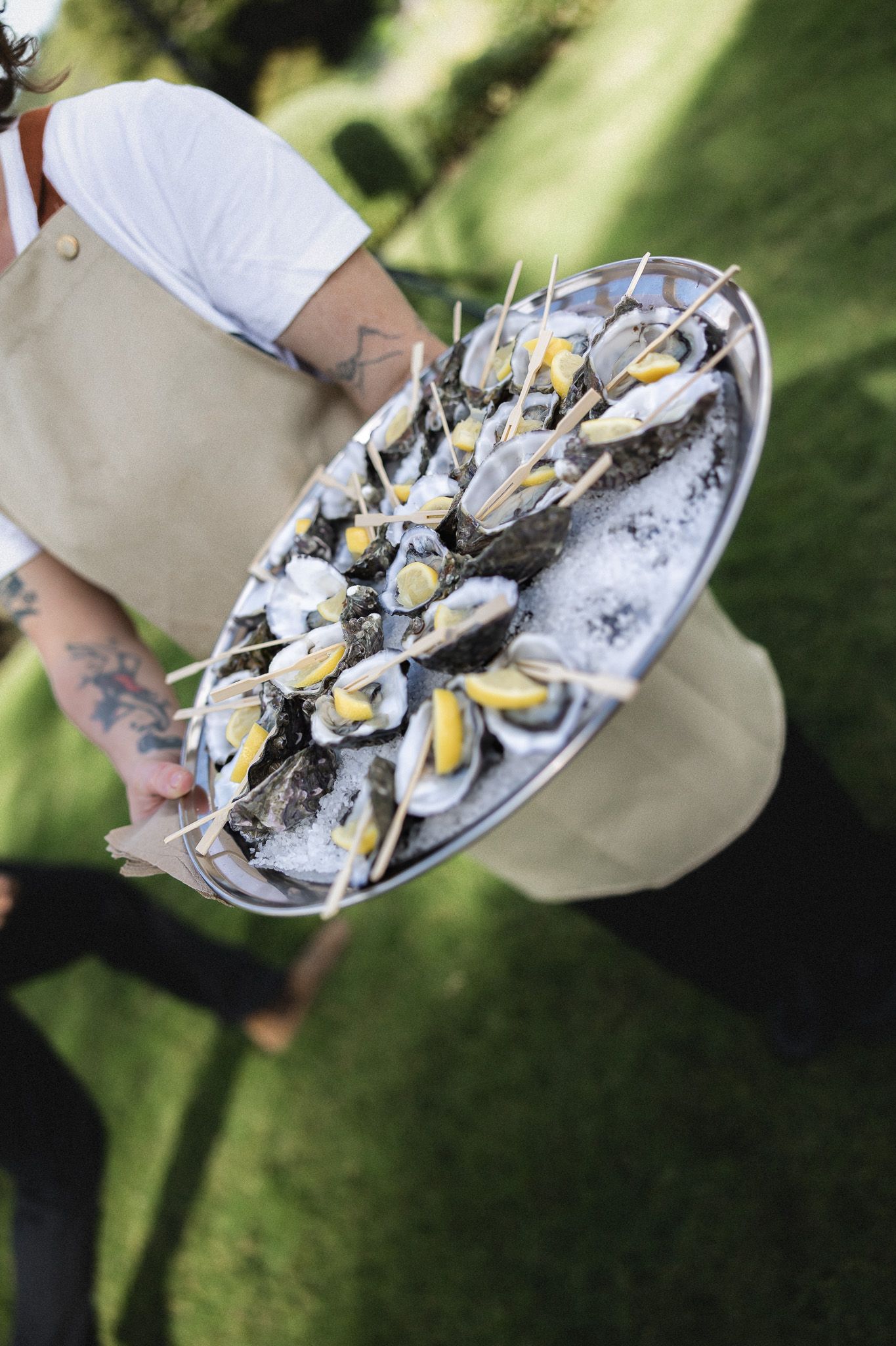 A tattooed server holds a tray of fresh oysters on ice at an outdoor wedding reception.
