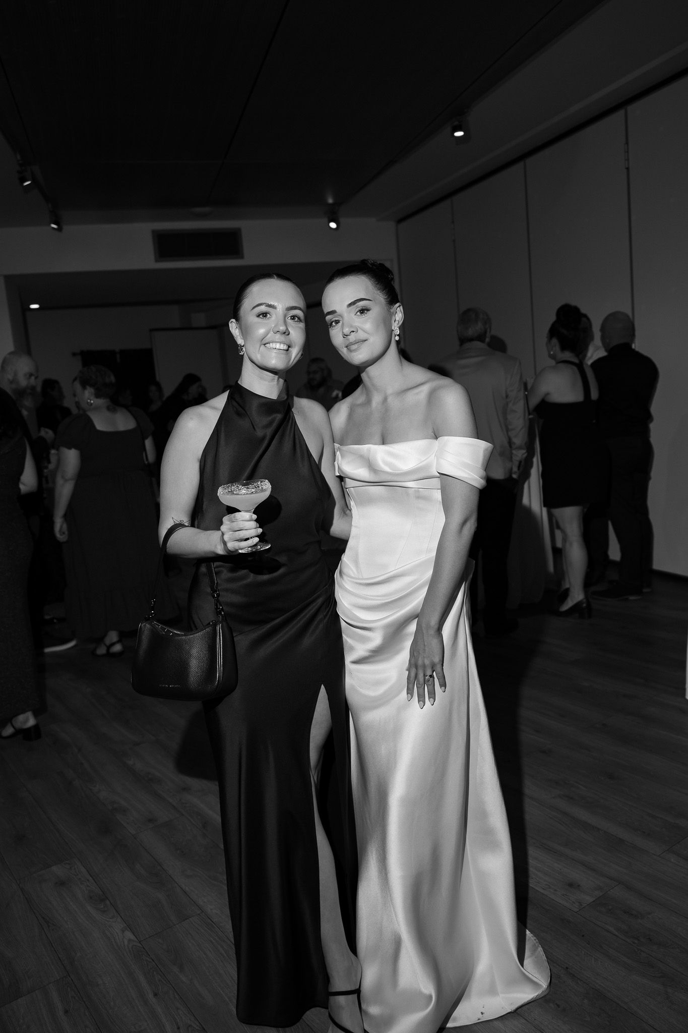 Two elegantly dressed women pose together at a wedding reception, one in black and one in white satin gown.