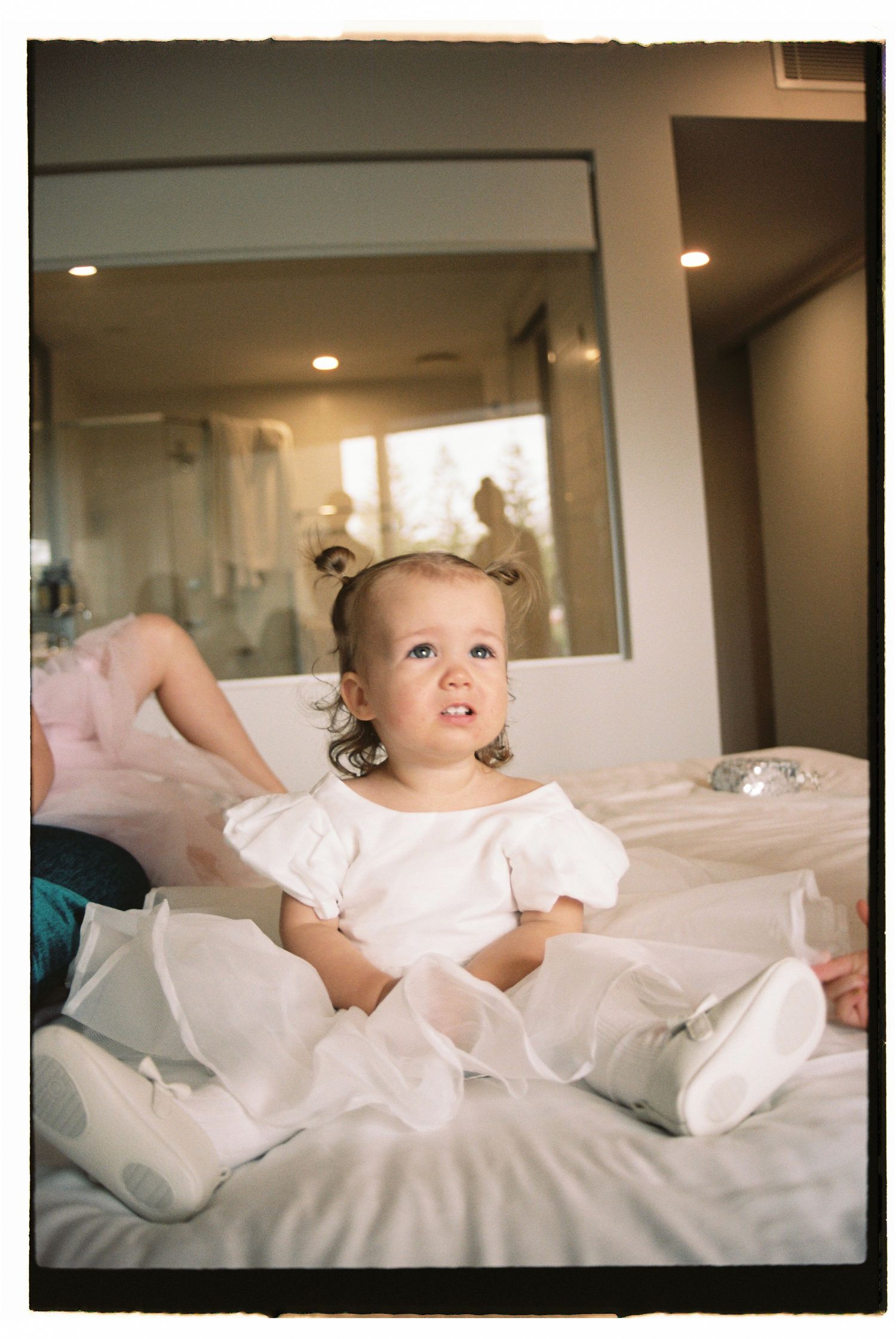 Adorable toddler girl in white dress with puff sleeves sits on bed with blue eyes gazing up.