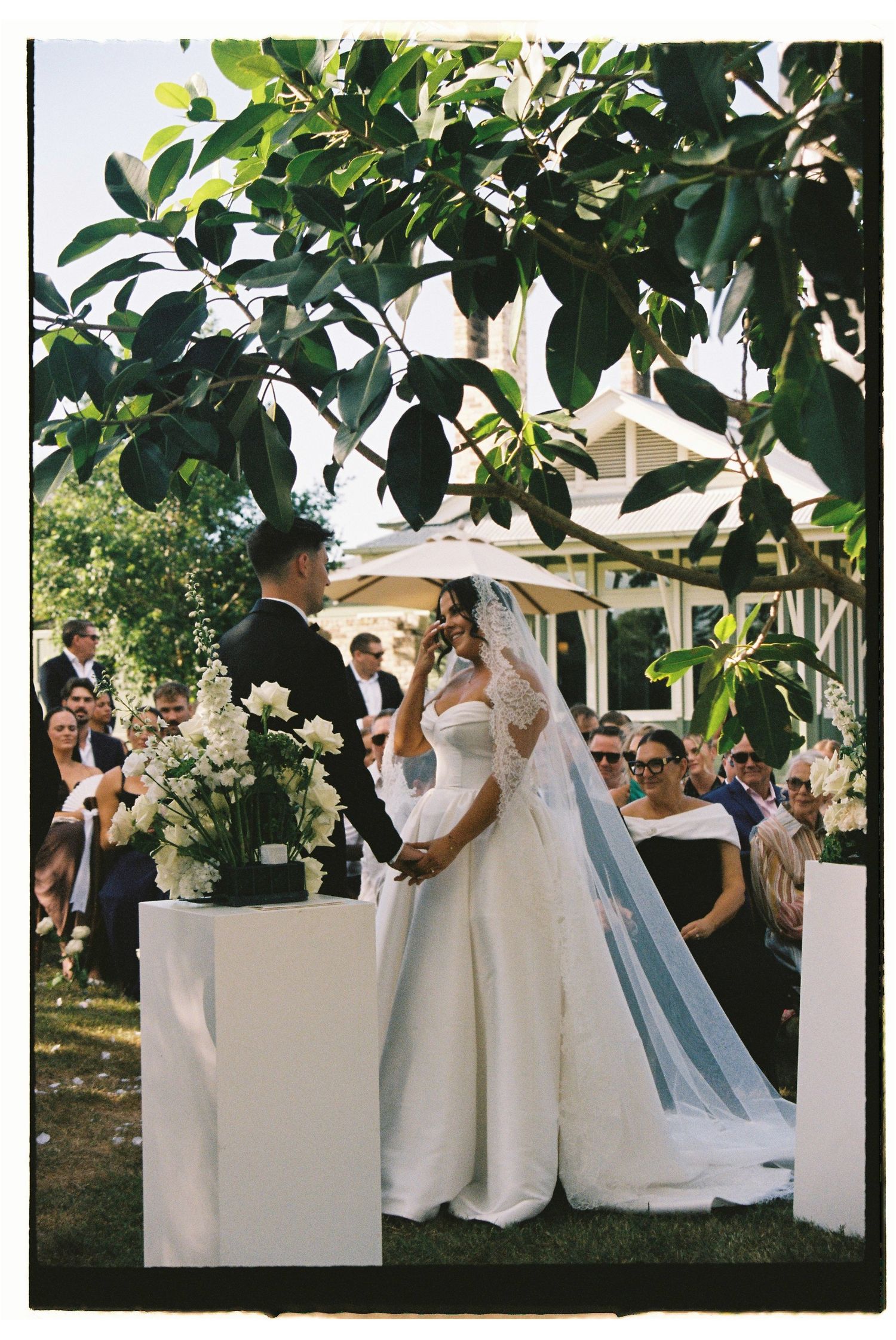 Bride in elegant off-shoulder gown with veil exchanges vows with groom at outdoor garden wedding ceremony.