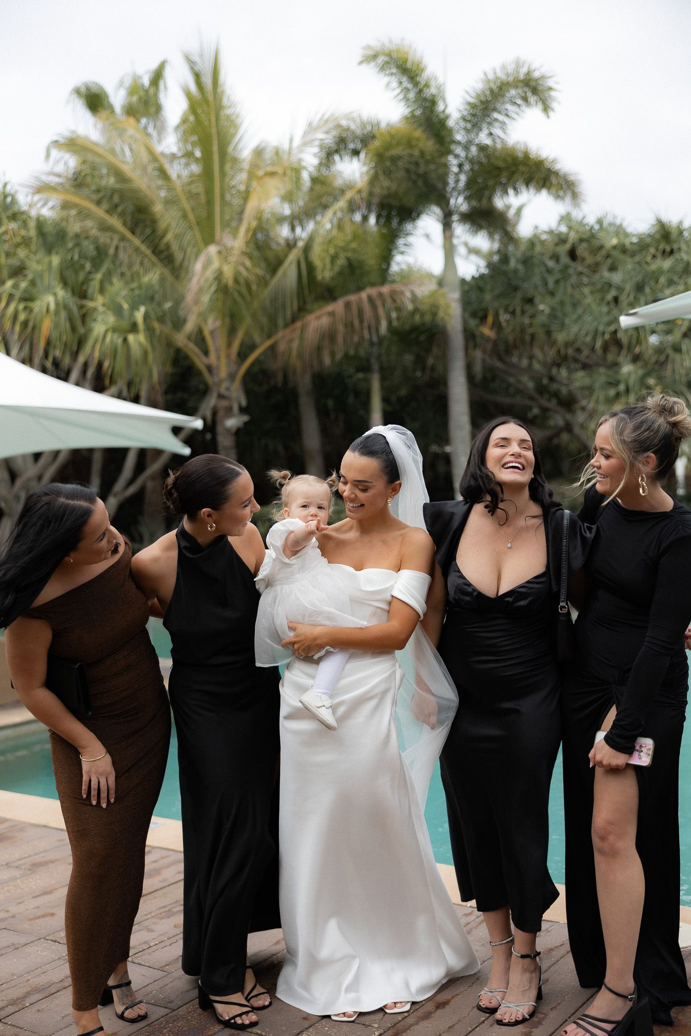 Bride in white gown holding baby surrounded by bridesmaids in black dresses near pool with palm trees.