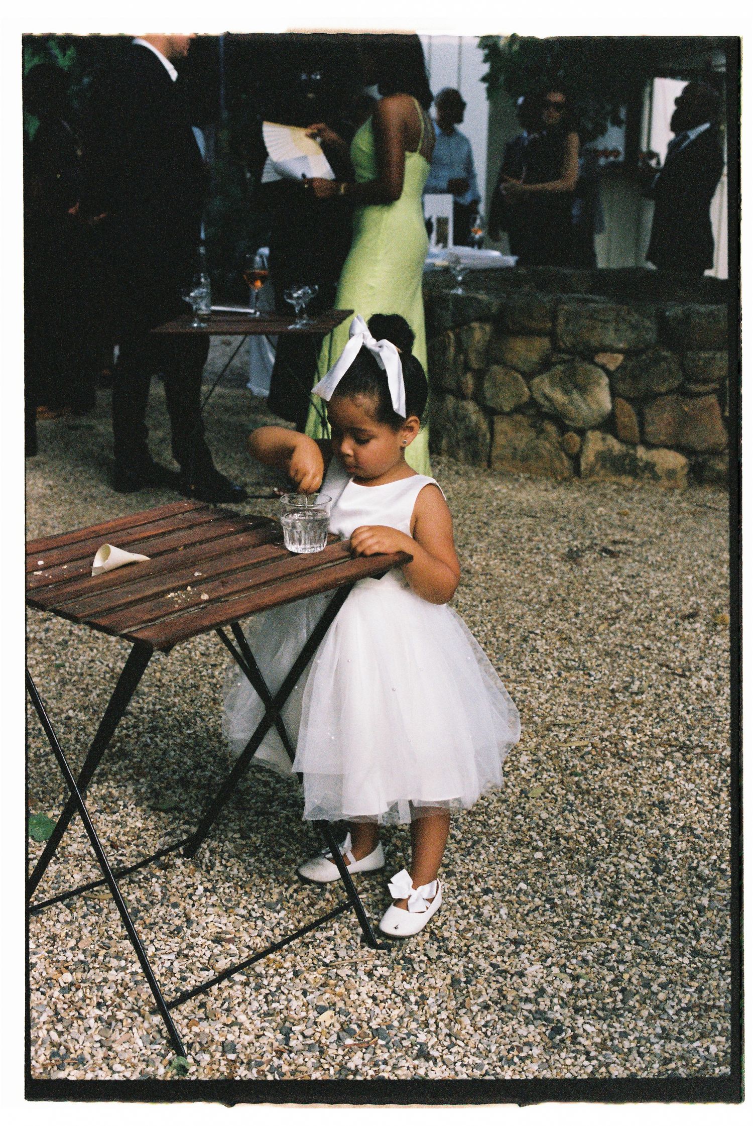 A young flower girl in a white tulle dress and bow headband stands at a wooden table during an outdoor wedding reception.