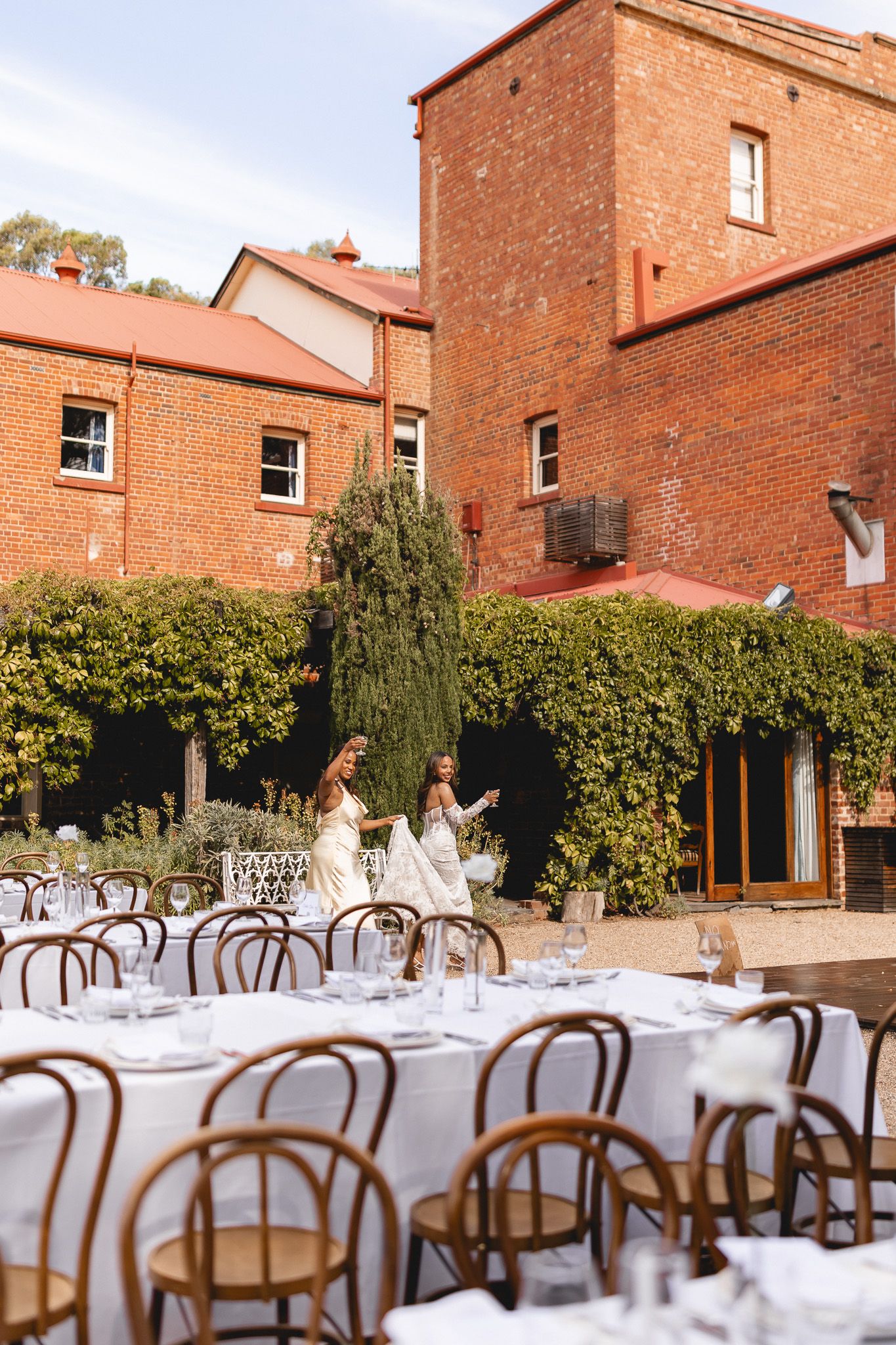 Outdoor wedding reception setup with round tables, bentwood chairs, and red brick heritage building backdrop.
