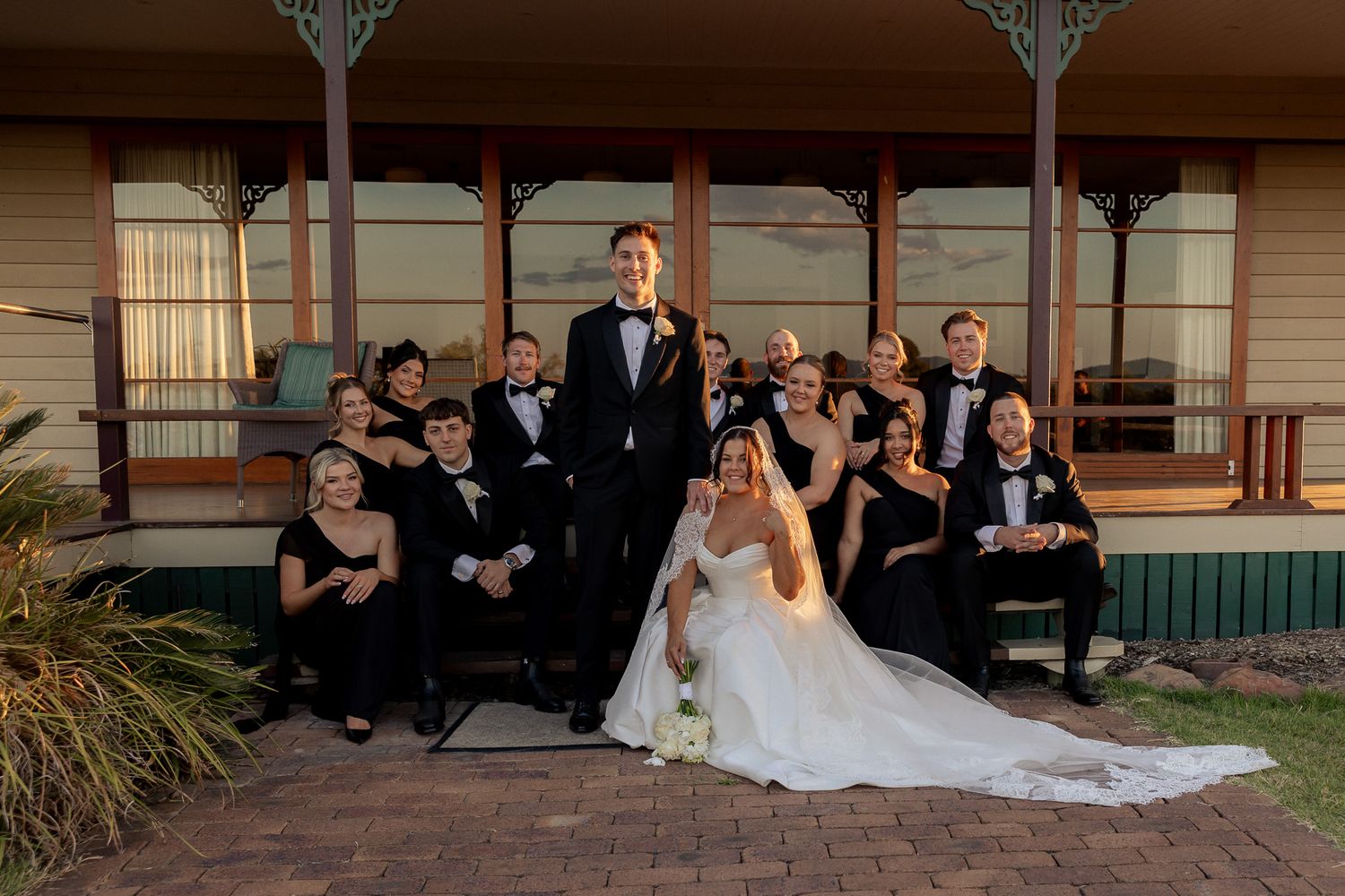 Wedding party poses on porch steps at golden hour, bride in white gown surrounded by guests in black formal attire.