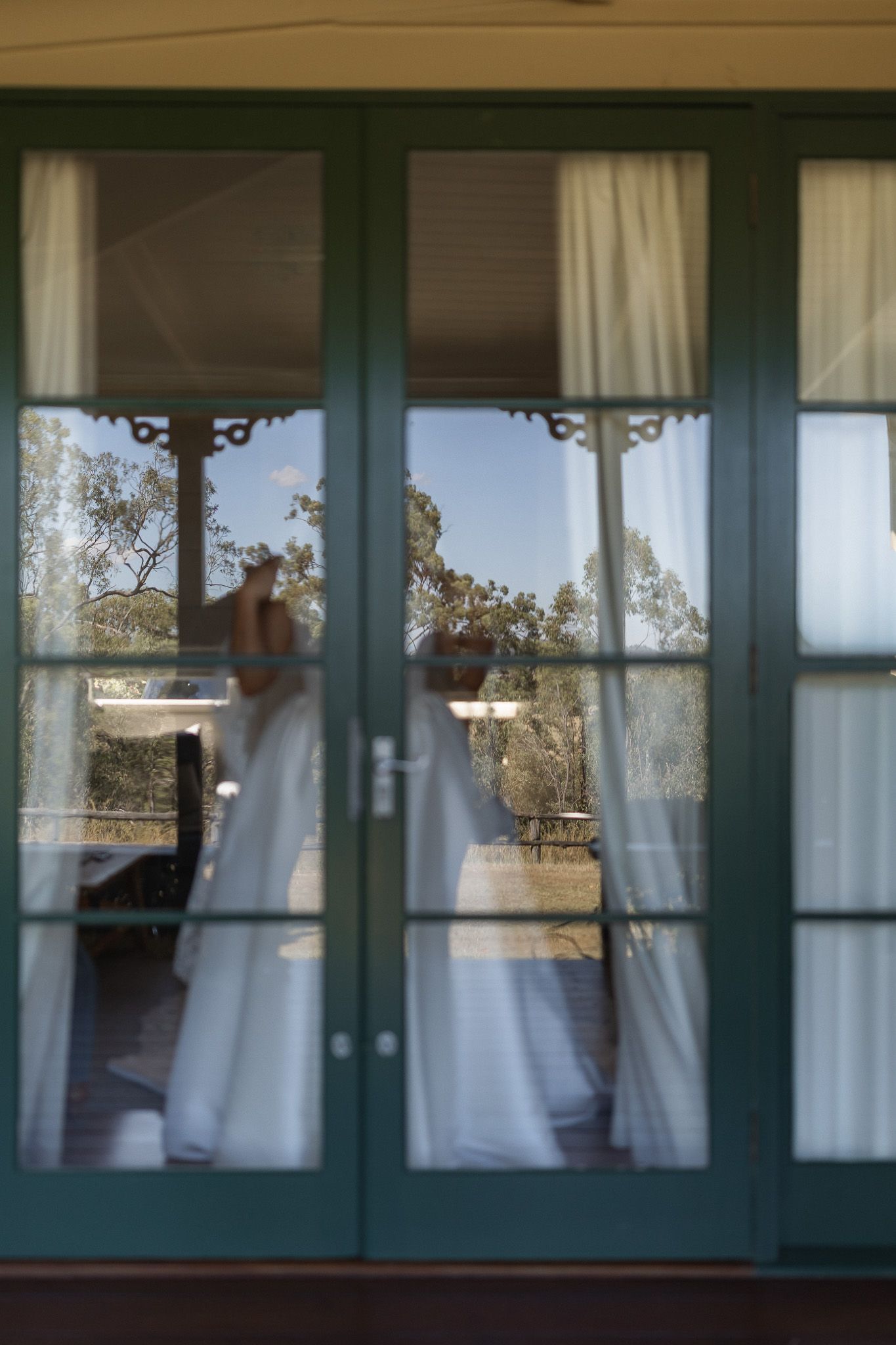 Bride in white wedding dress seen through green-framed French doors with sheer curtains and outdoor trees in background.