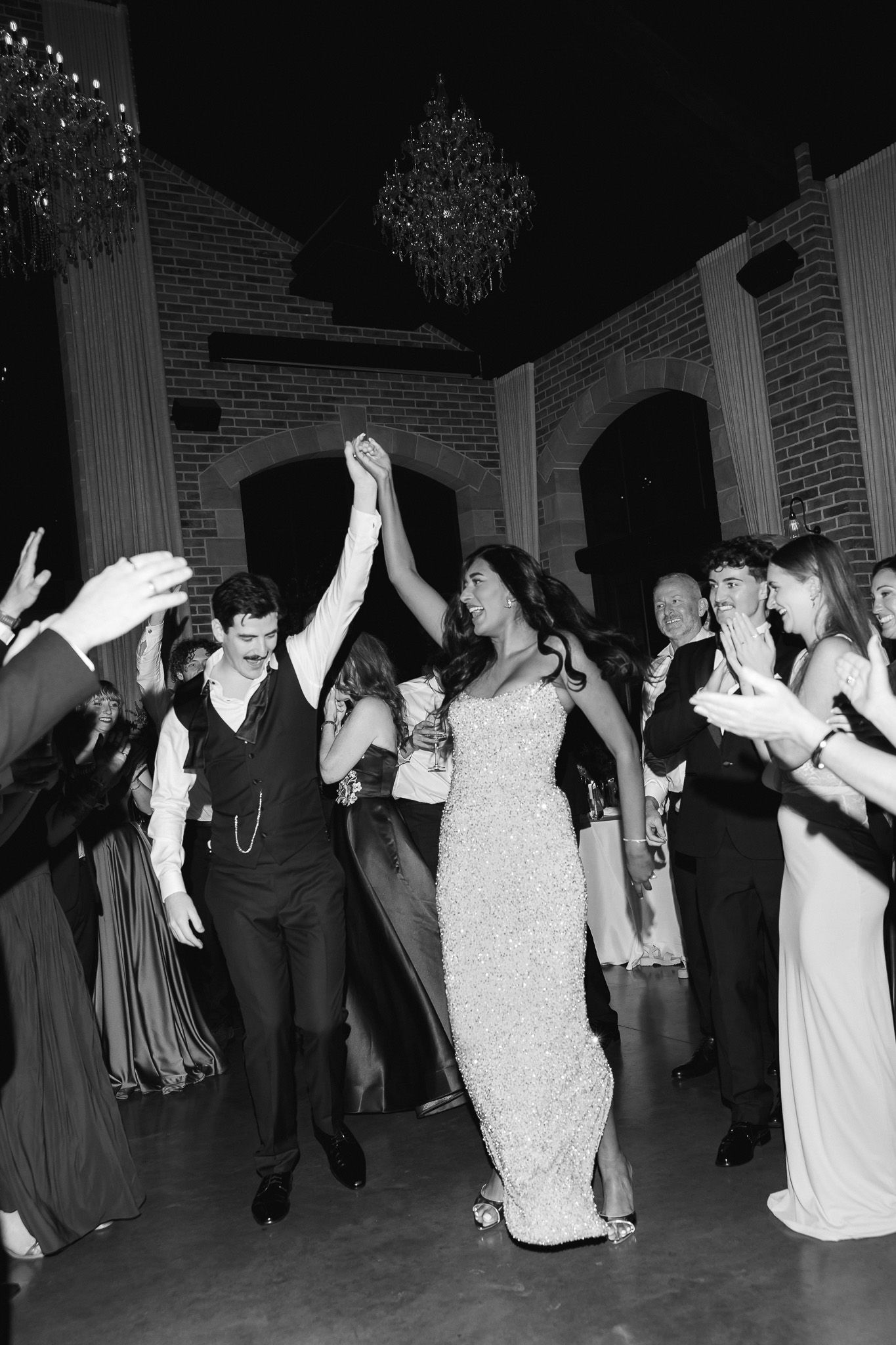 Black and white photo of bride and groom dancing joyfully with guests at elegant wedding reception.