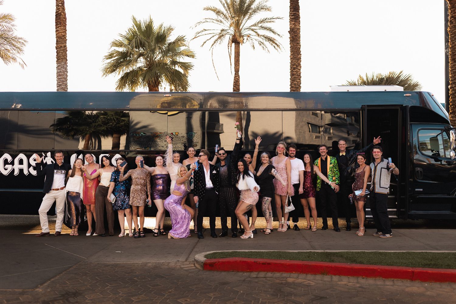 Large group of elegantly dressed wedding guests posing in front of a party bus surrounded by palm trees.