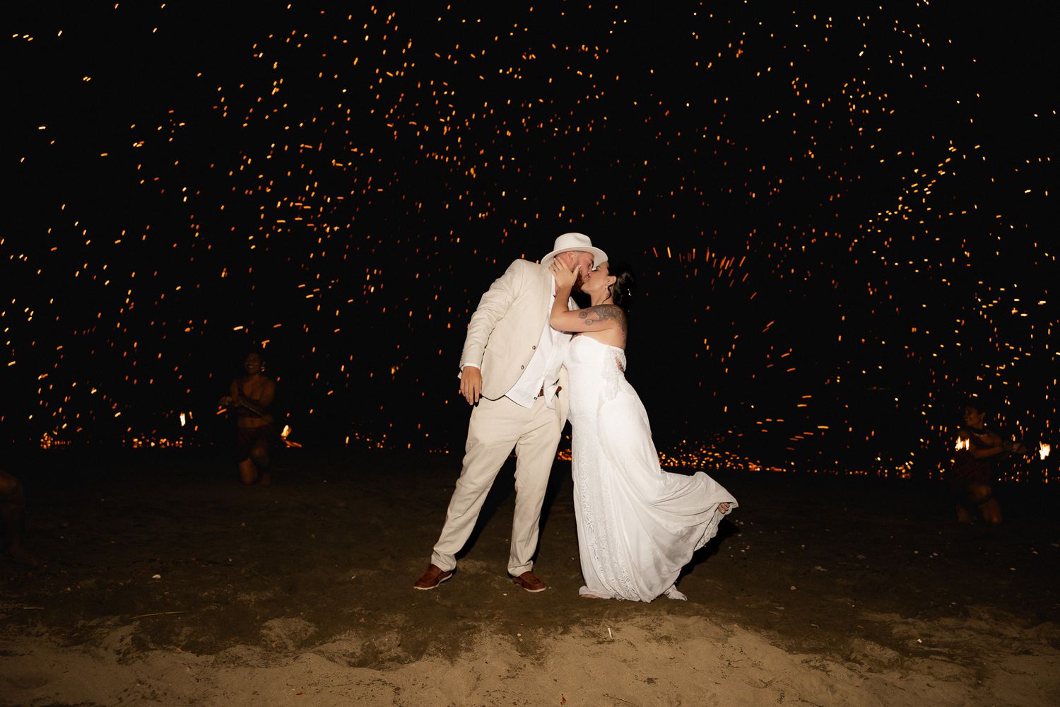 Bride and groom share romantic kiss surrounded by sparkling fireworks display on beach at night.