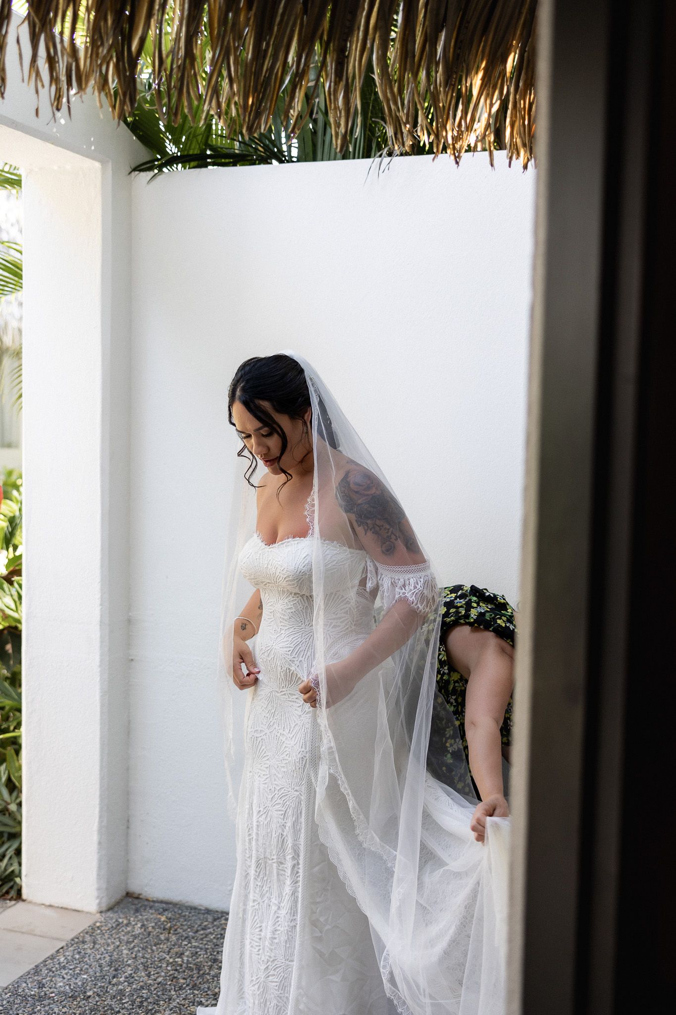 Bride in off-shoulder lace wedding gown and long veil stands outdoors near white wall with tropical thatched roof.