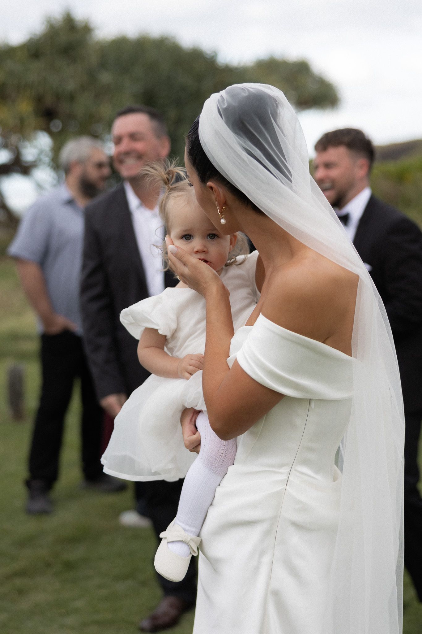 Bride in white gown and veil tenderly kisses baby girl during outdoor wedding ceremony.