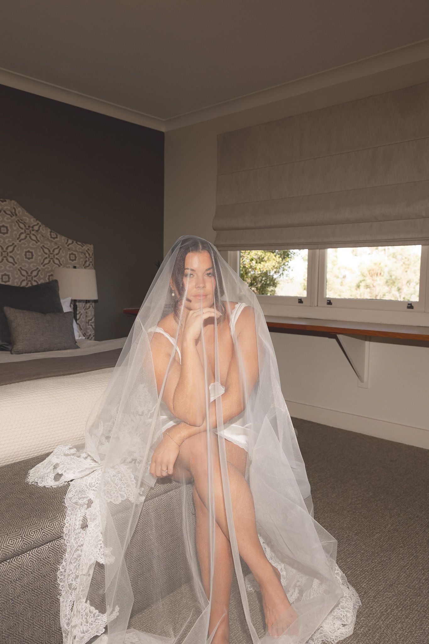 Bride draped in lace-trimmed cathedral veil sits gracefully in lingerie in a softly lit bedroom.