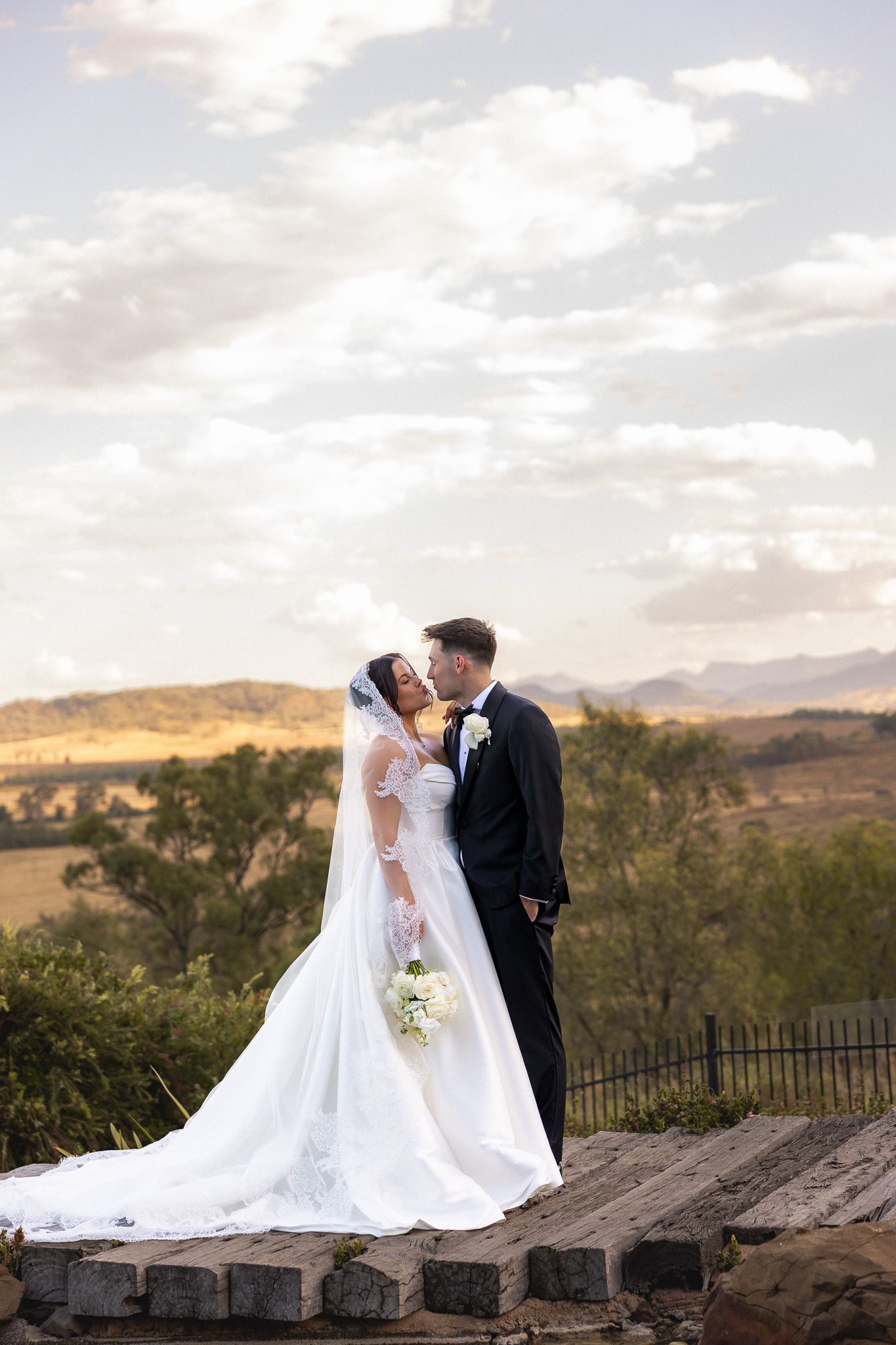 Bride and groom sharing a romantic kiss on a wooden platform overlooking a scenic countryside landscape at sunset.
