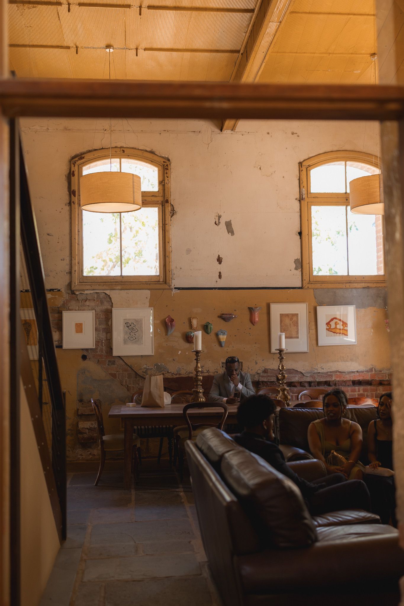 Rustic interior lounge with leather sofas, artwork on distressed walls, pendant lamps, and guests relaxing near tall arched windows.